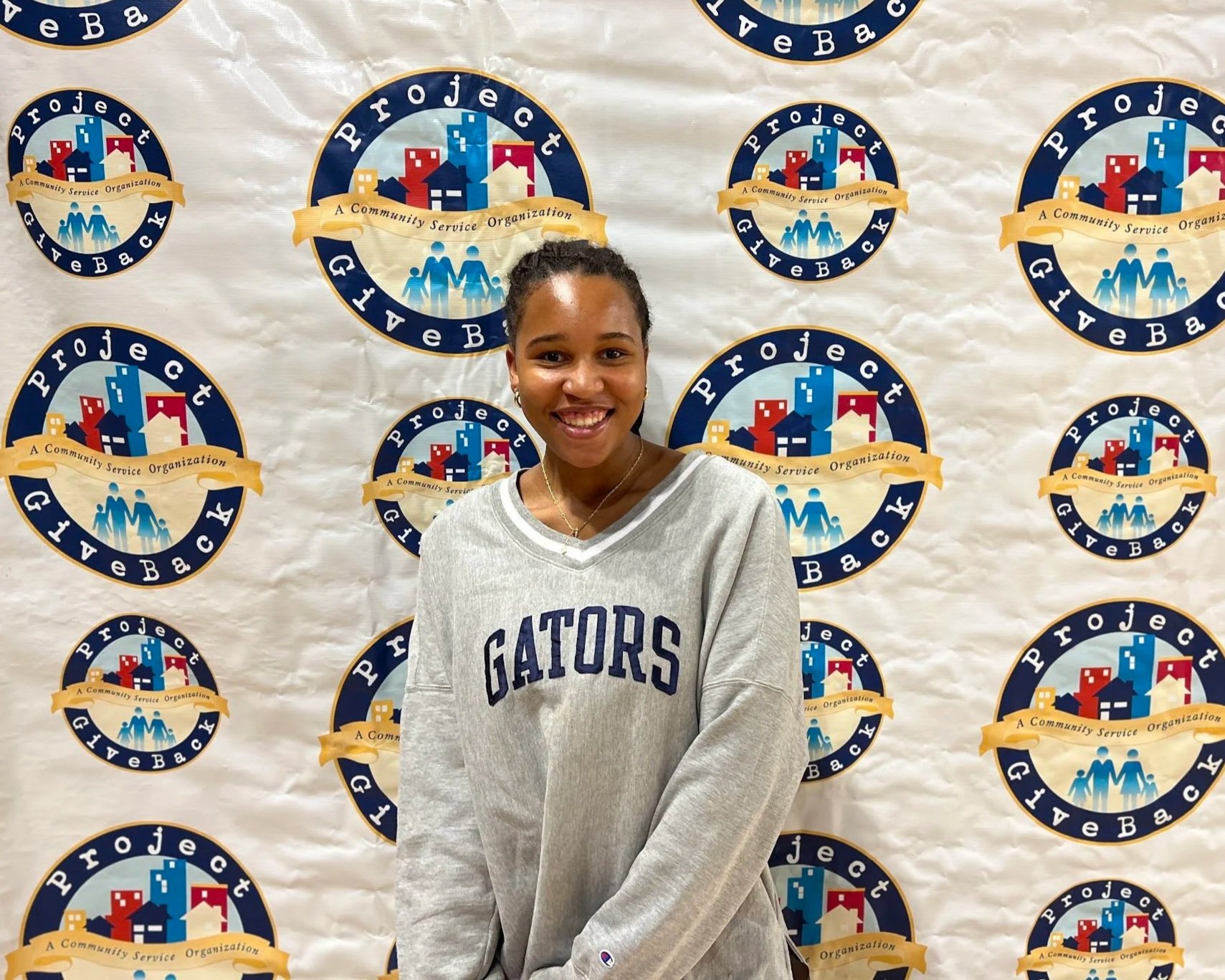 A young woman smiling in front of a backdrop with multiple logos of a community service organization. She is wearing a gray Gators sweatshirt.