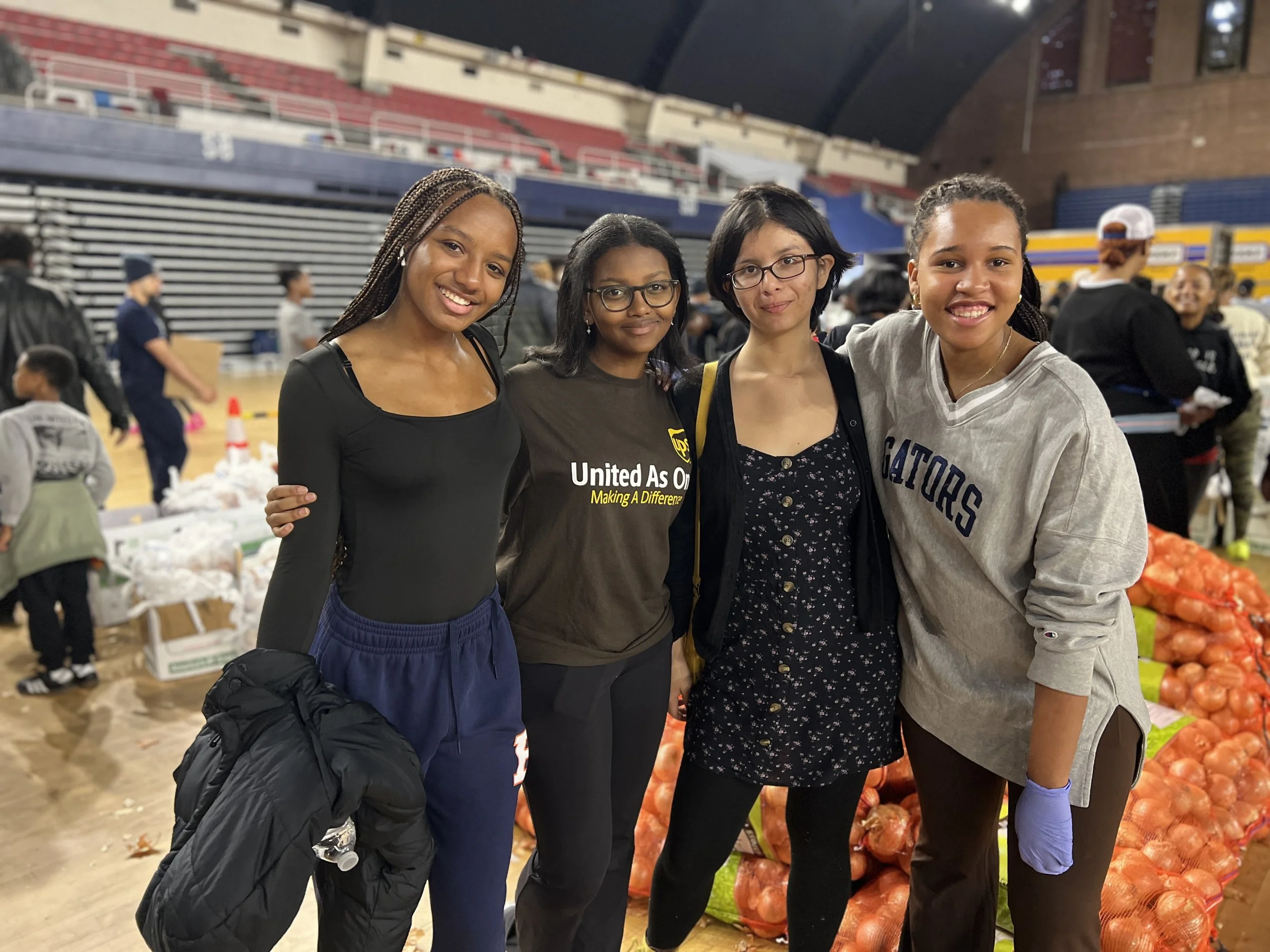 Four young women stand together at an indoor charity event, smiling at the camera. They are surrounded by bags of onions and other food items, with volunteers and people in the background.