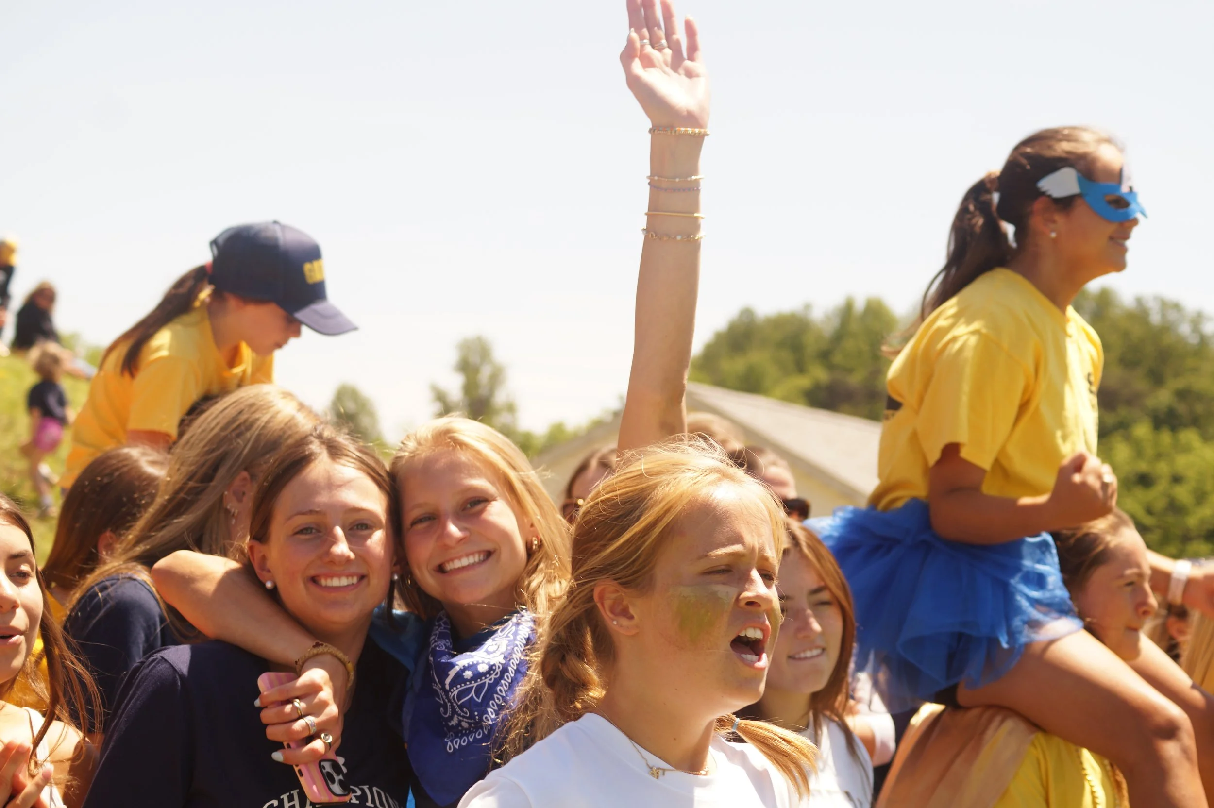 A group of smiling young women, some with face paint and wearing bandanas, are gathered outdoors on a sunny day. They are closely packed, celebrating or participating in an event. One woman in a yellow shirt and blue mask is on someone's shoulders, raising her arm. The background shows trees and a clear blue sky.