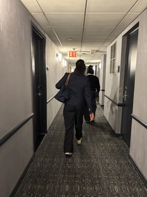 Two women walk down a dimly lit hotel hallway with dark doors on either side, an exit sign visible at the end.