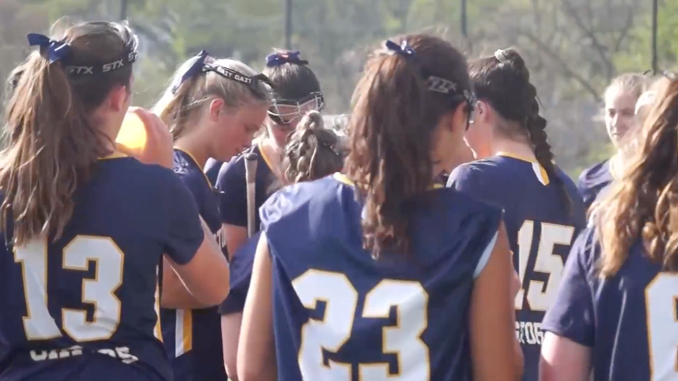A group of female athletes in blue jerseys with yellow accents and numbers, some with braided hair, gathered outdoors on a sunny day, possibly during a sports practice or game.