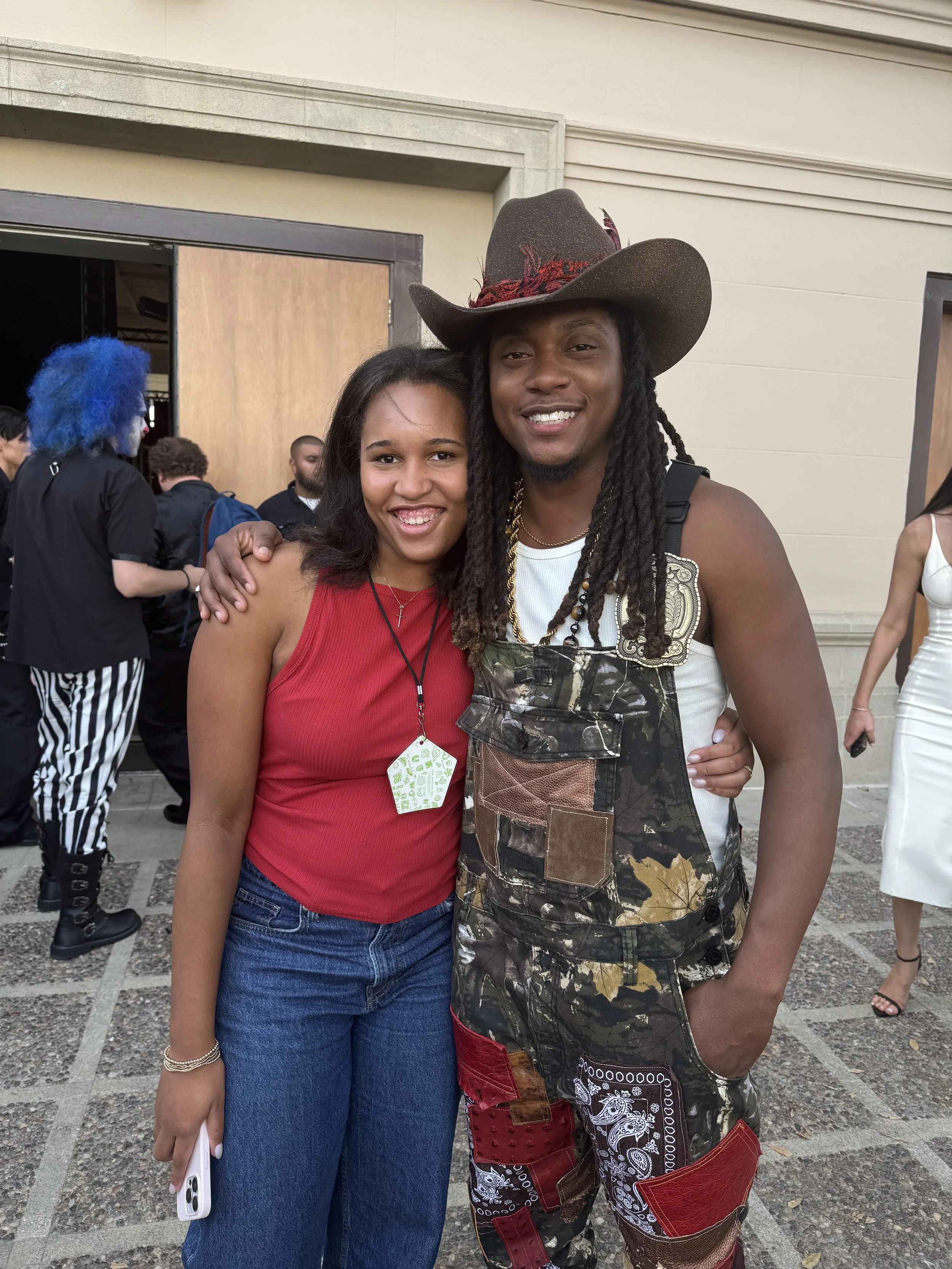 A young woman and a man with long dreadlocks, one wearing a red top and the other wearing a wide-brimmed hat and overalls, smiling and standing close together outside at an event with other people in the background.