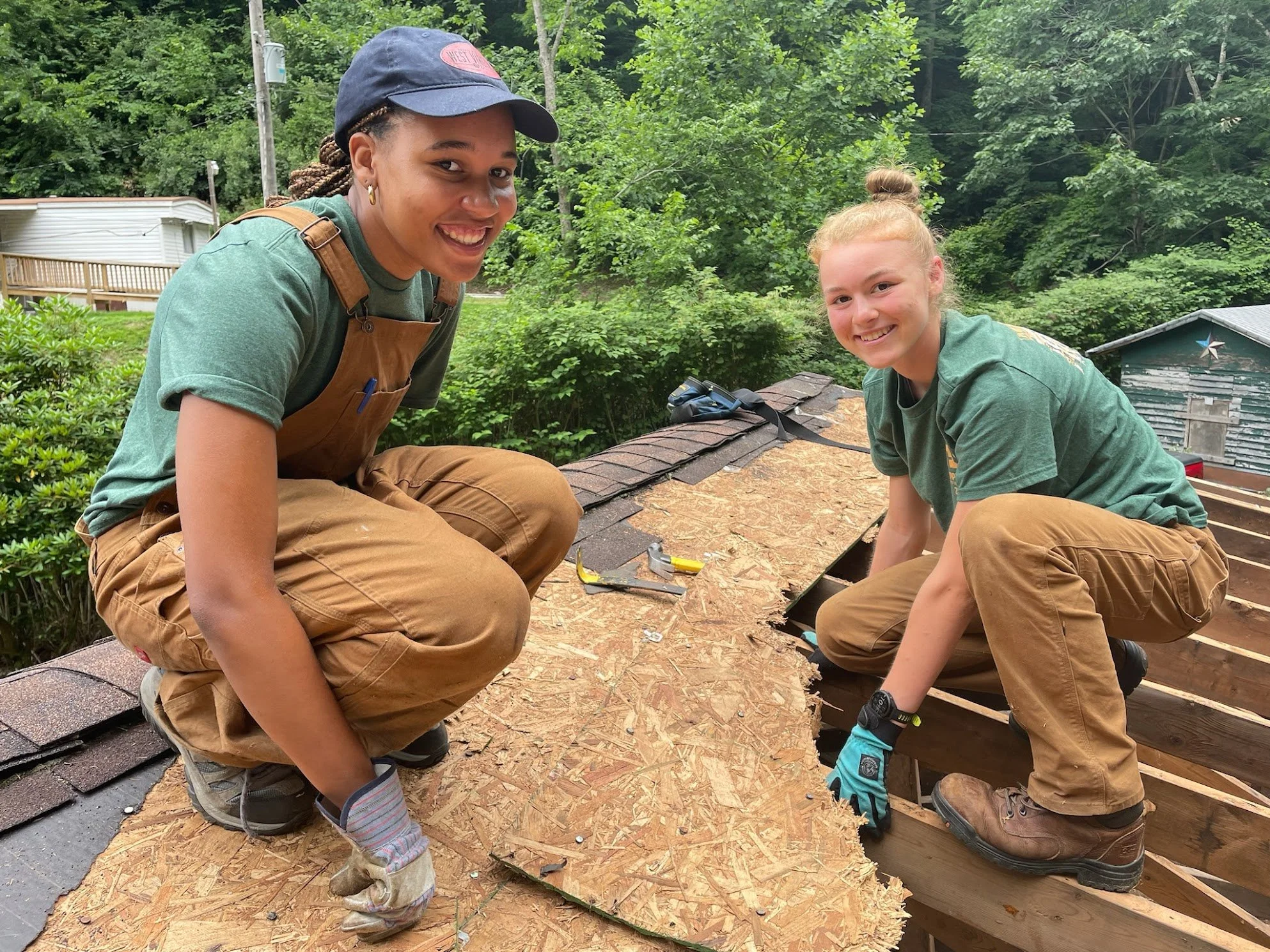 Two women working on a roofing project outdoors surrounded by green trees, kneeling on a wooden roof frame, with tools nearby and smiling at the camera.