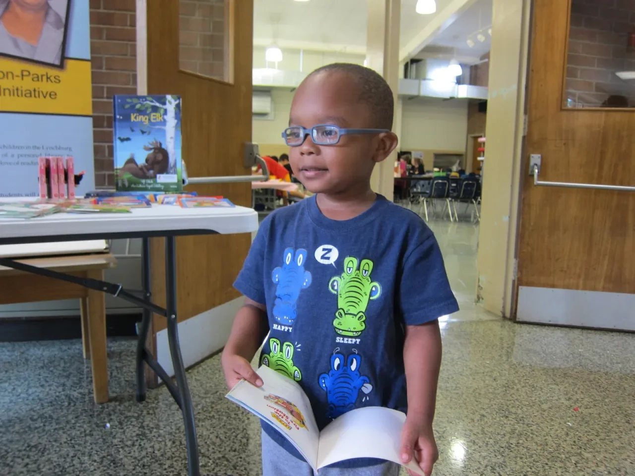 A young boy wearing glasses and a blue shirt with cartoon crocodiles and hippos, holding a book, standing in a room with tables, posters, and other children in the background.