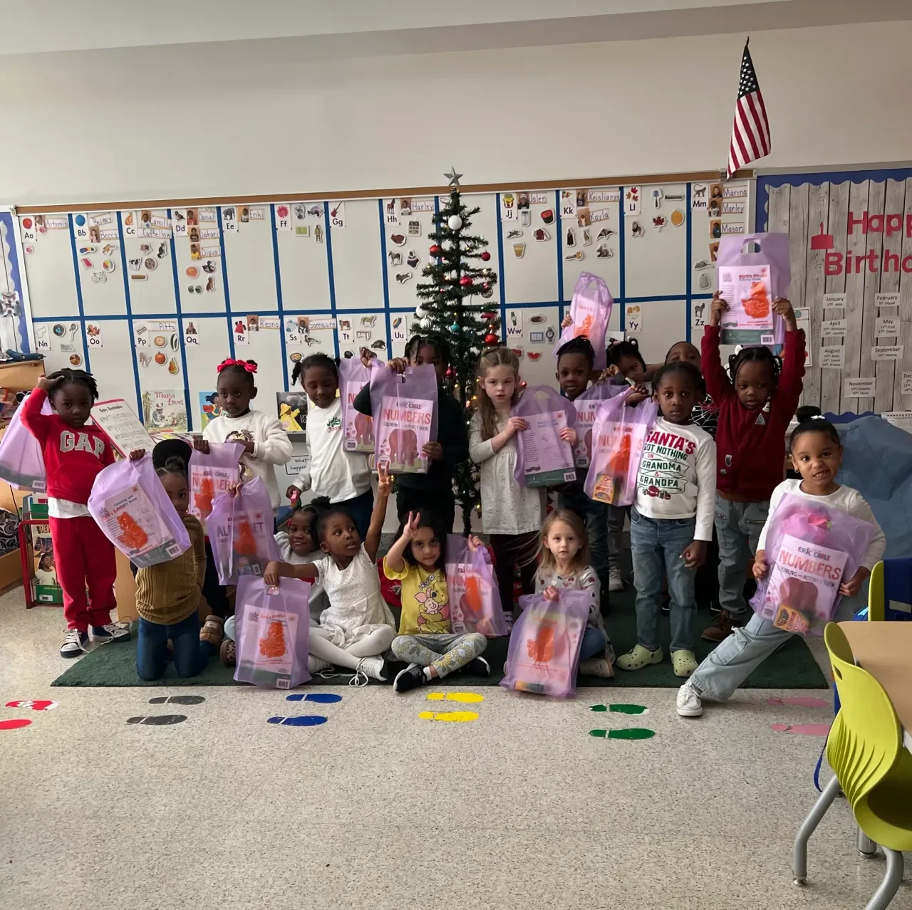 Group of children in a classroom, holding pink gift bags, with a Christmas tree and educational posters in the background.
