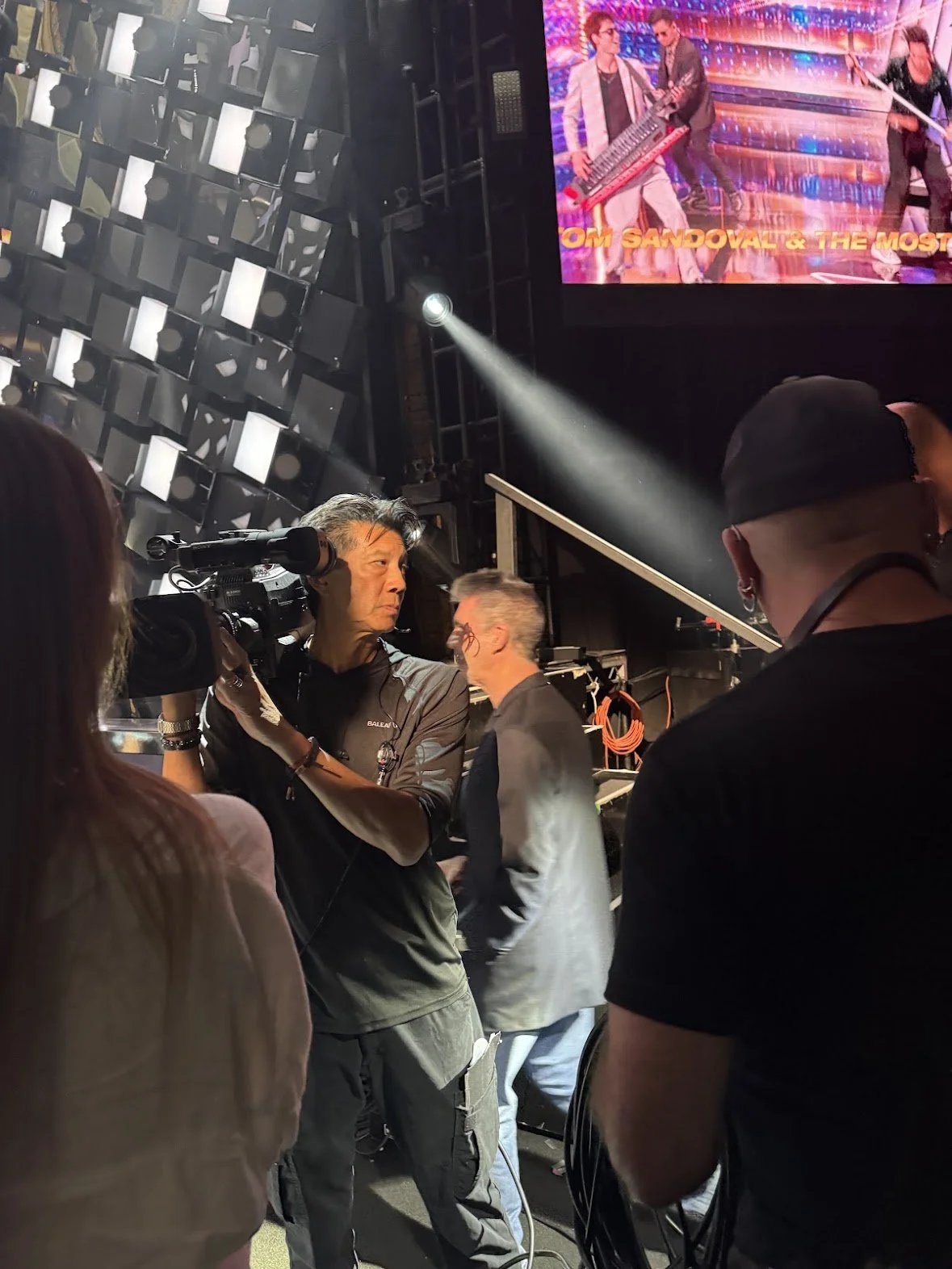 People working behind the scenes at a concert or event, with a large screen displaying performers in the background.