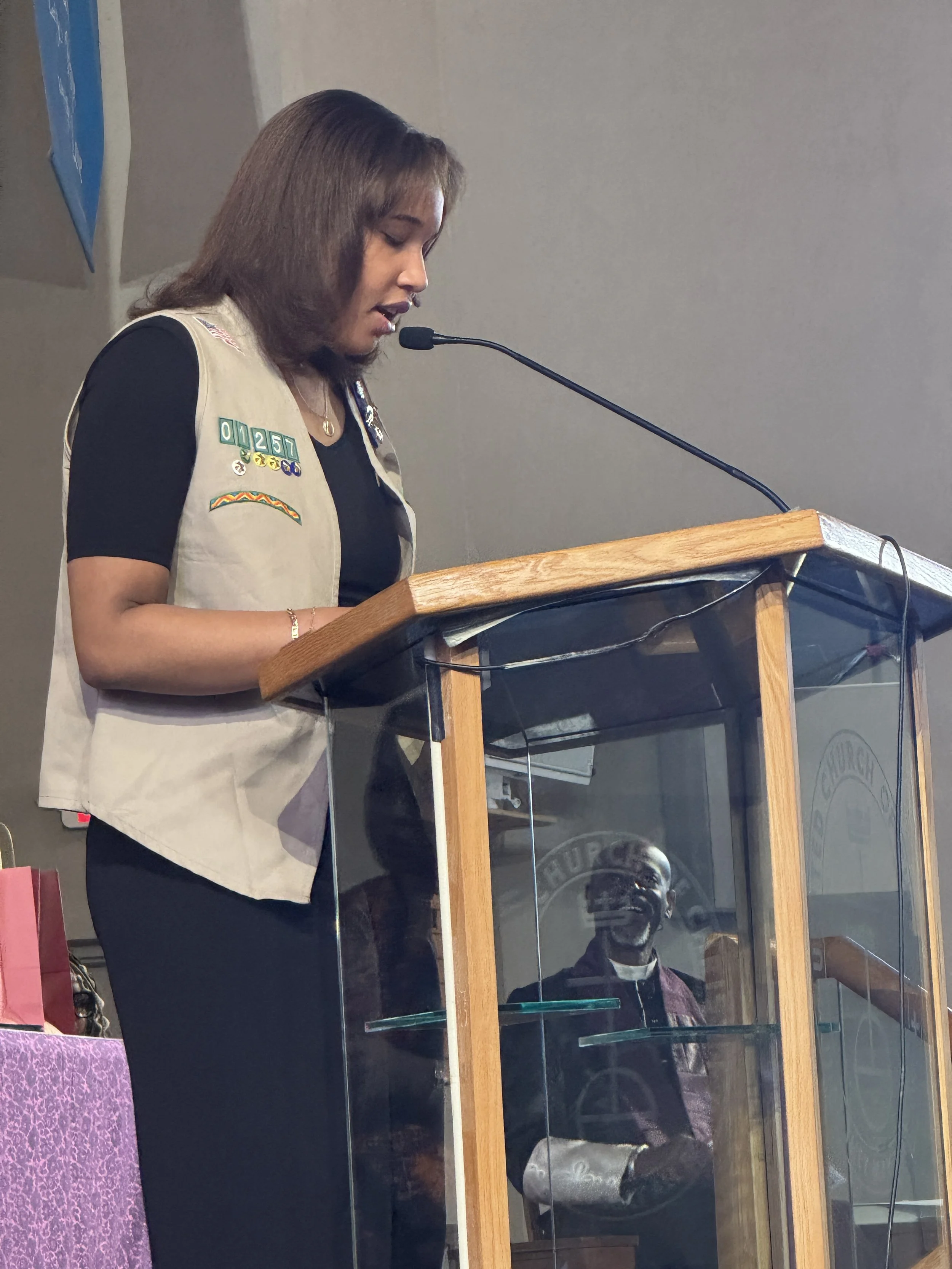 Woman wearing a beige vest and black shirt speaking at a podium with a microphone, with a man dressed in dark clothing and glasses reflected in the glass panel below.