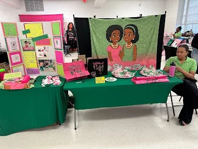 A table with pink and black items, including a cartoon backdrop of two girls, in a classroom setting with students and display boards.