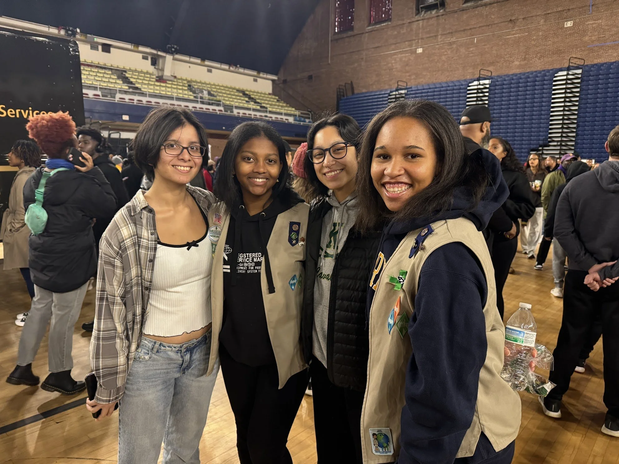 Four young women stand close together, smiling at the camera inside a crowded gymnasium or event hall. The women appear to be diverse in ethnicity, and are dressed in casual and scout uniforms. The background shows other people, a brick wall, and rows of yellow and blue seating, indicating a social or community event.