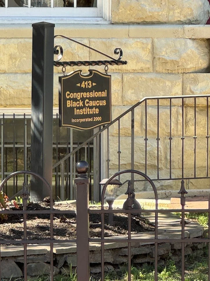 Black and yellow sign for Congressional Black Caucus Institute hanging outside a stone building, with a small garden and metal fence in front.