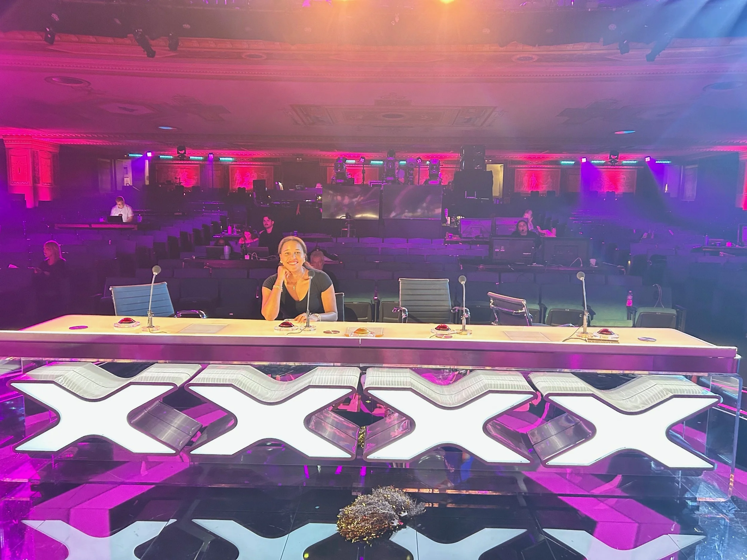 A woman sitting at a table with microphones in a conference room with colorful stage lighting and empty chairs and tables.