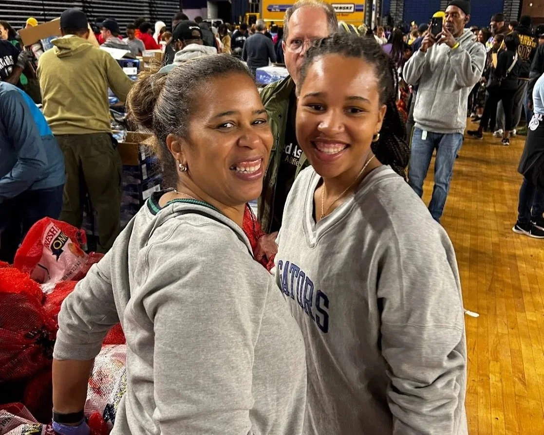 Two women smiling at the camera in a crowded indoor event space, with many people in the background, some taking photos and others working at tables.