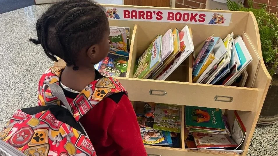 A young girl with braided hair wearing a red jacket and carrying a backpack with a game controller pattern, looking at a wooden book display labeled 'Barb's Books' filled with children's books at a library or bookstore.