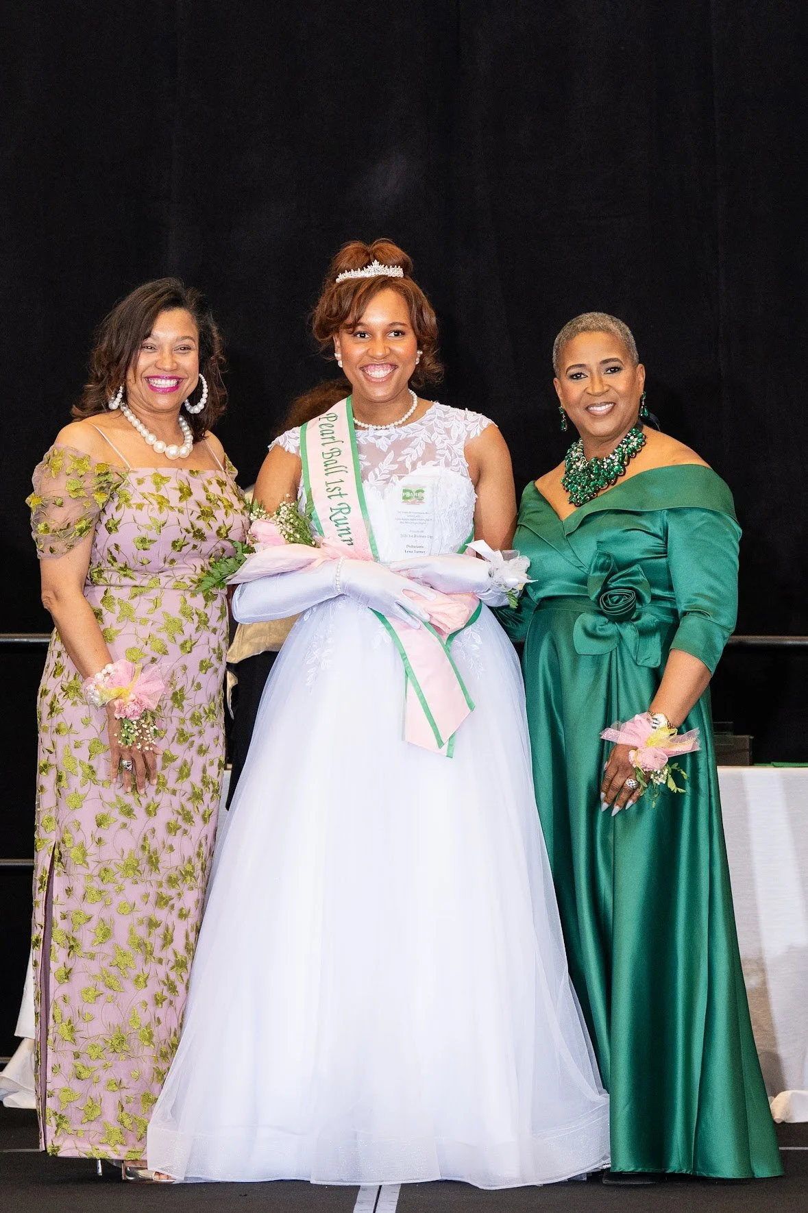 Three women in formal attire standing on stage, one in a pink floral dress, another in a green gown, and the woman in the middle in a white dress with a sash and crown, holding a plaque.