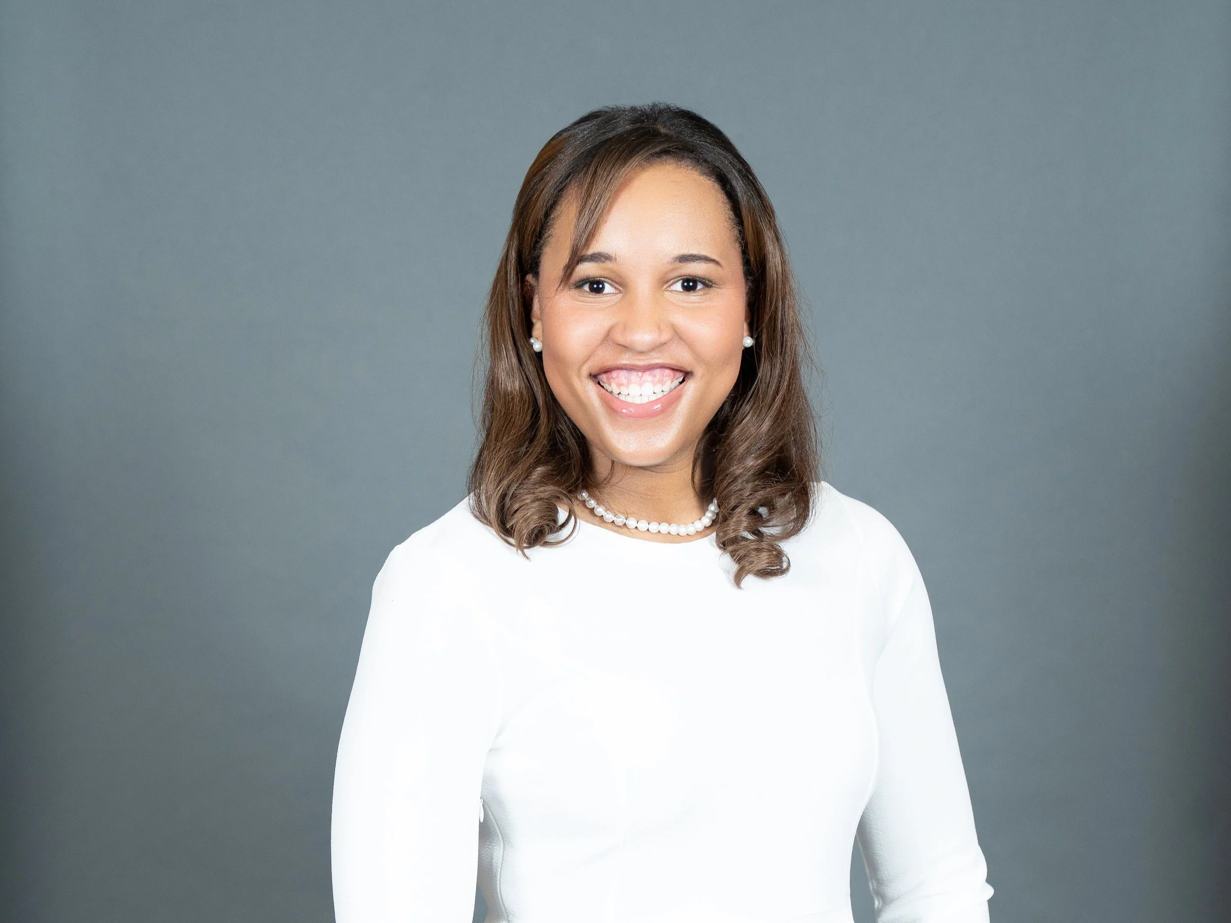 A woman smiling with long brown hair, wearing pearl earrings, a pearl necklace, and a white top, standing against a plain gray background.
