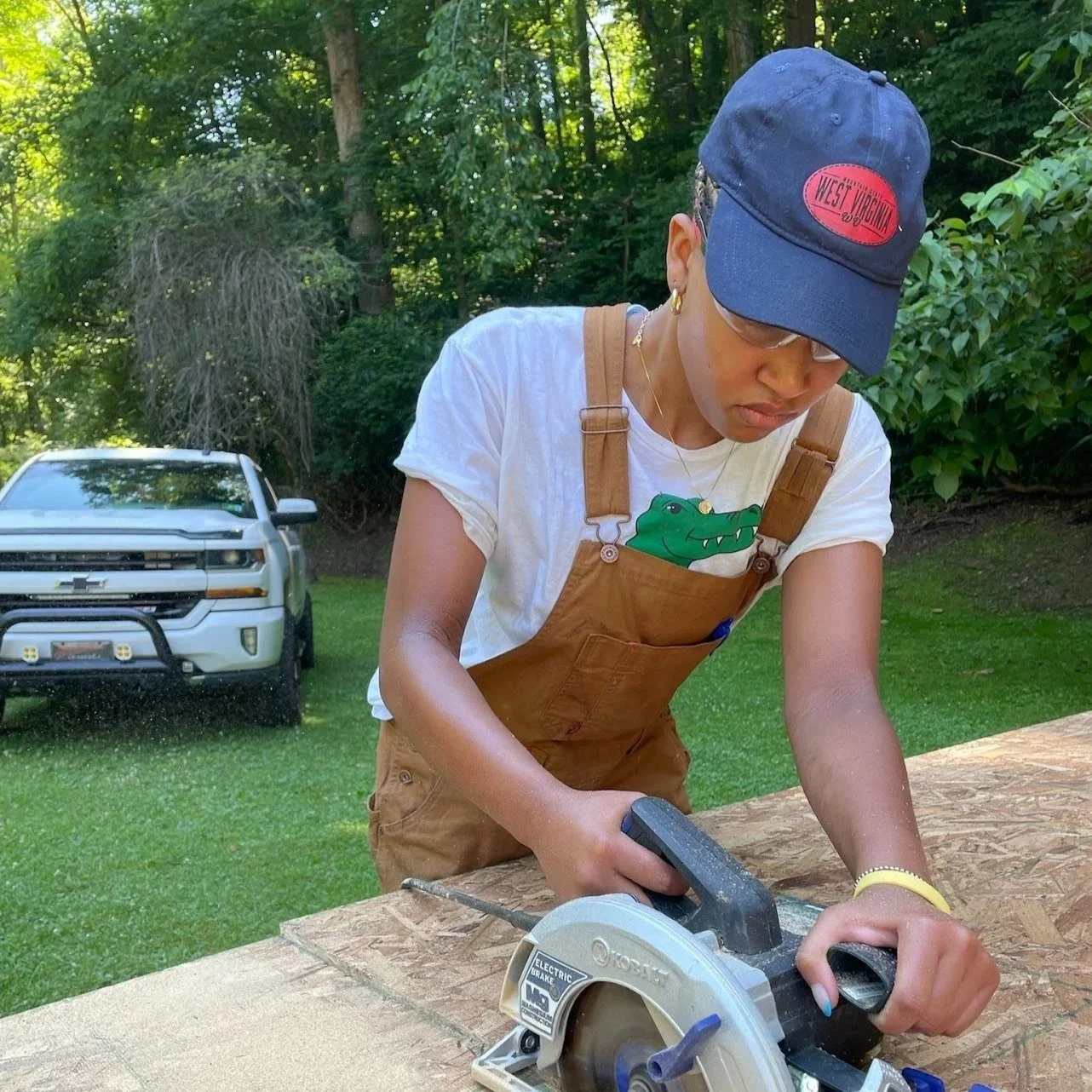 A woman wearing a blue cap, glasses, and a white T-shirt with a crocodile graphic works with a circular saw at a woodworking project outdoors on a wooden surface, with a green grassy yard and trees, and a white vehicle in the background.