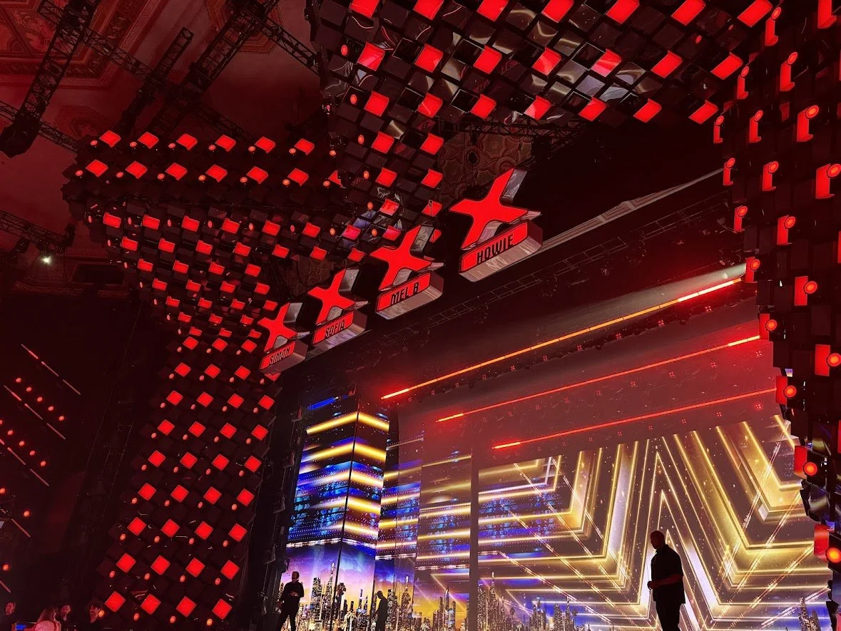 Stage with large LED screen displaying city skyline and light patterns, surrounded by red illuminated cubic decorations and hanging signs labeled 'Simon,' 'Sobe,' 'Mel B,' and 'Howie.'