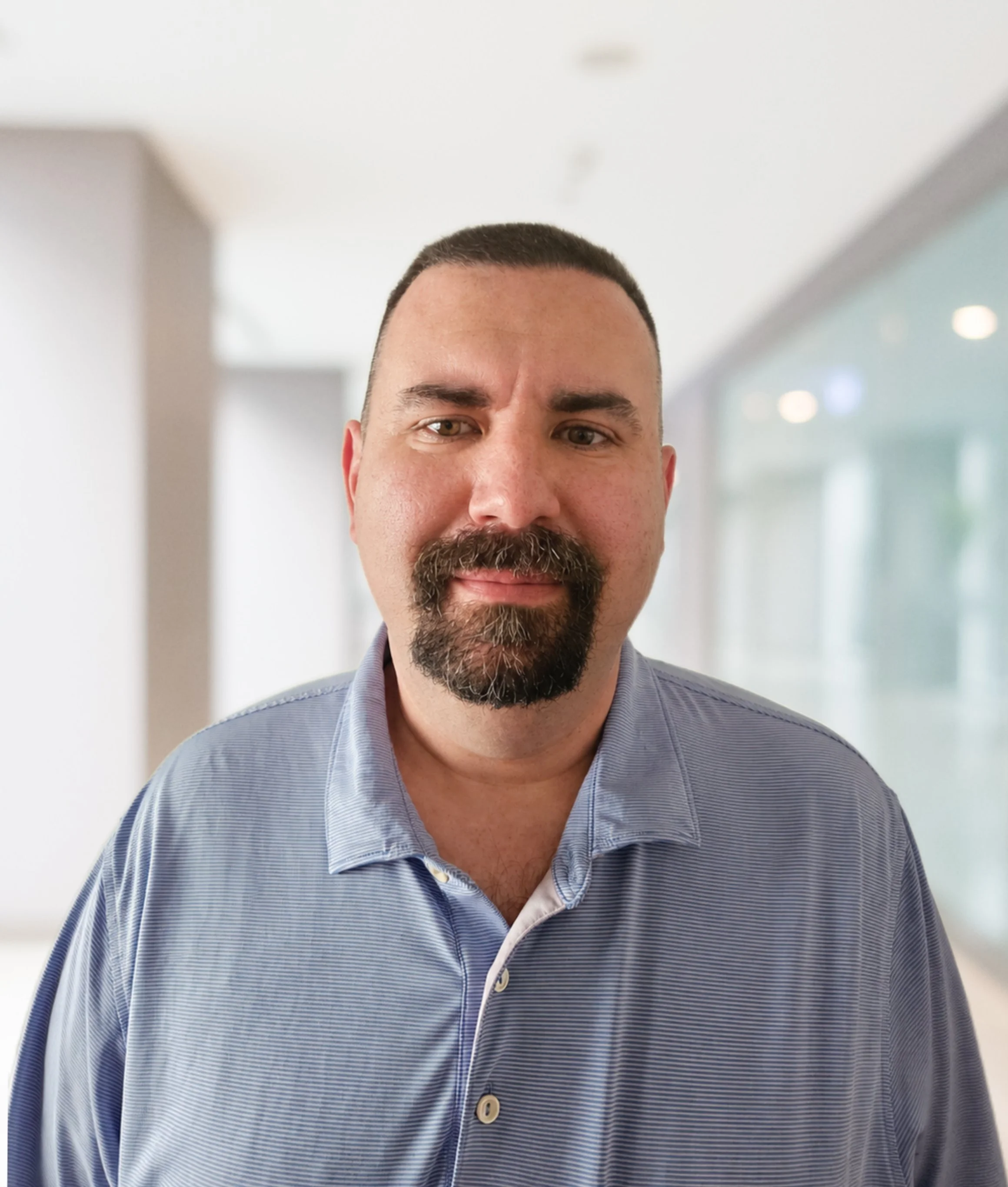 Headshot of Greg Stoerger, a man with short dark hair, a beard, and hazel eyes, wearing a light blue and white striped dress shirt with the top button undone, standing indoors with a blurred background.