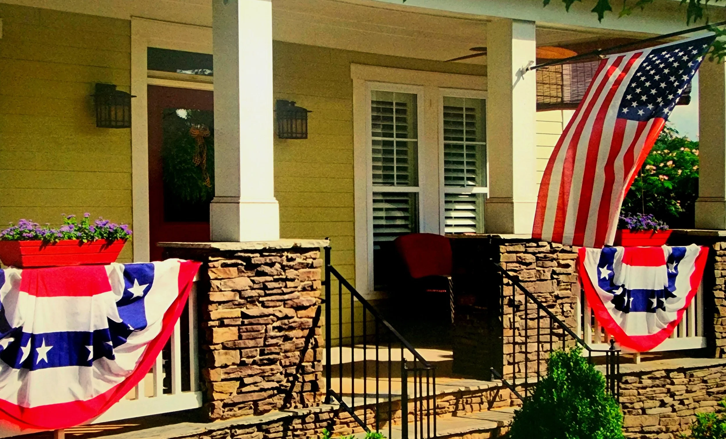 Front porch of a yellow house decorated for a patriotic celebration with red, white, and blue bunting, and an American flag hanging from the porch ceiling. The porch has a stone railing with flowers in red planters, and a porch light on each side of the door.