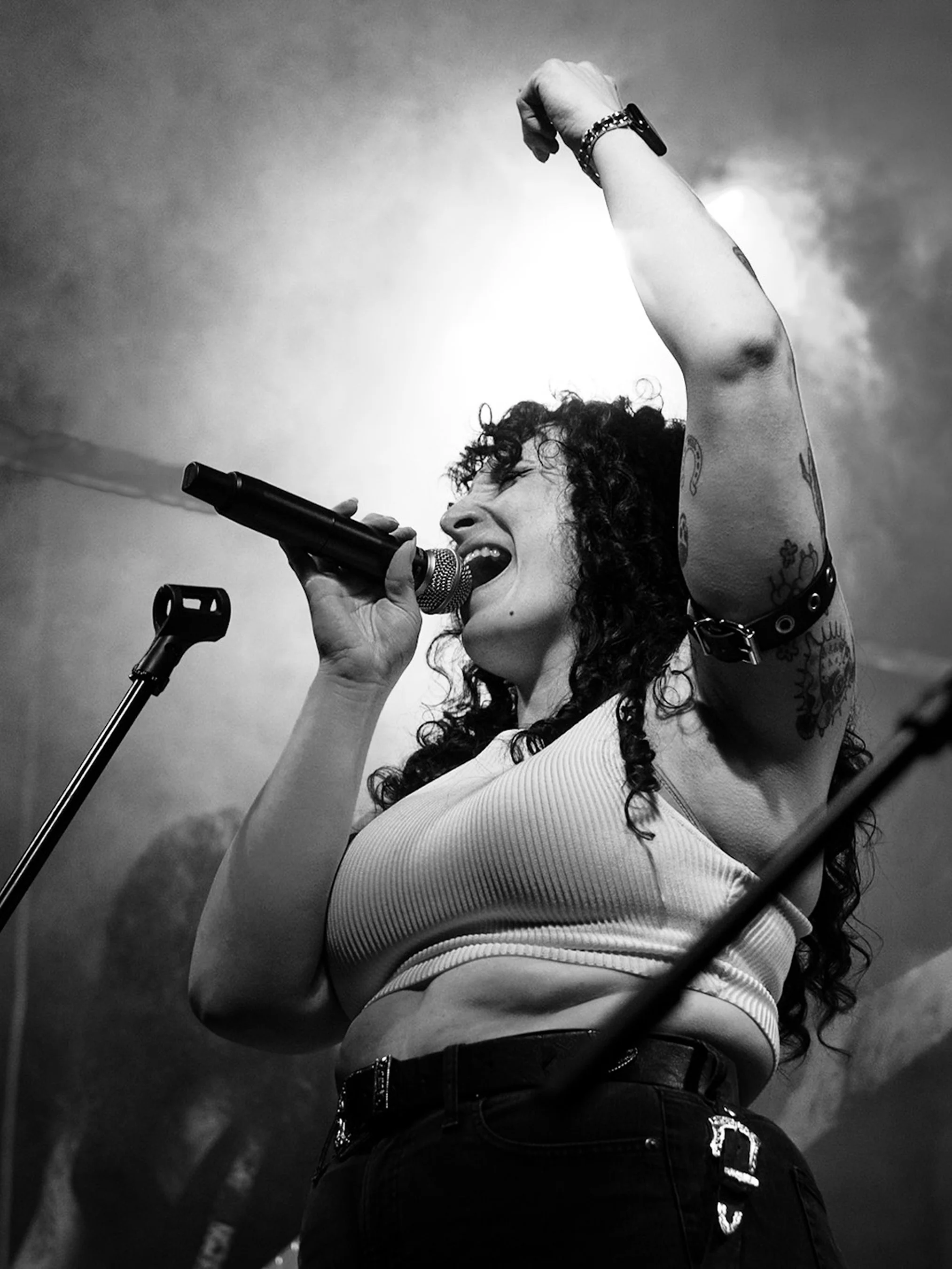 A woman singing into a microphone with her right arm raised and fist clenched, wearing a sleeveless top, watch, and bracelets, with tattoos on her arm, standing outdoors against a cloudy sky.
