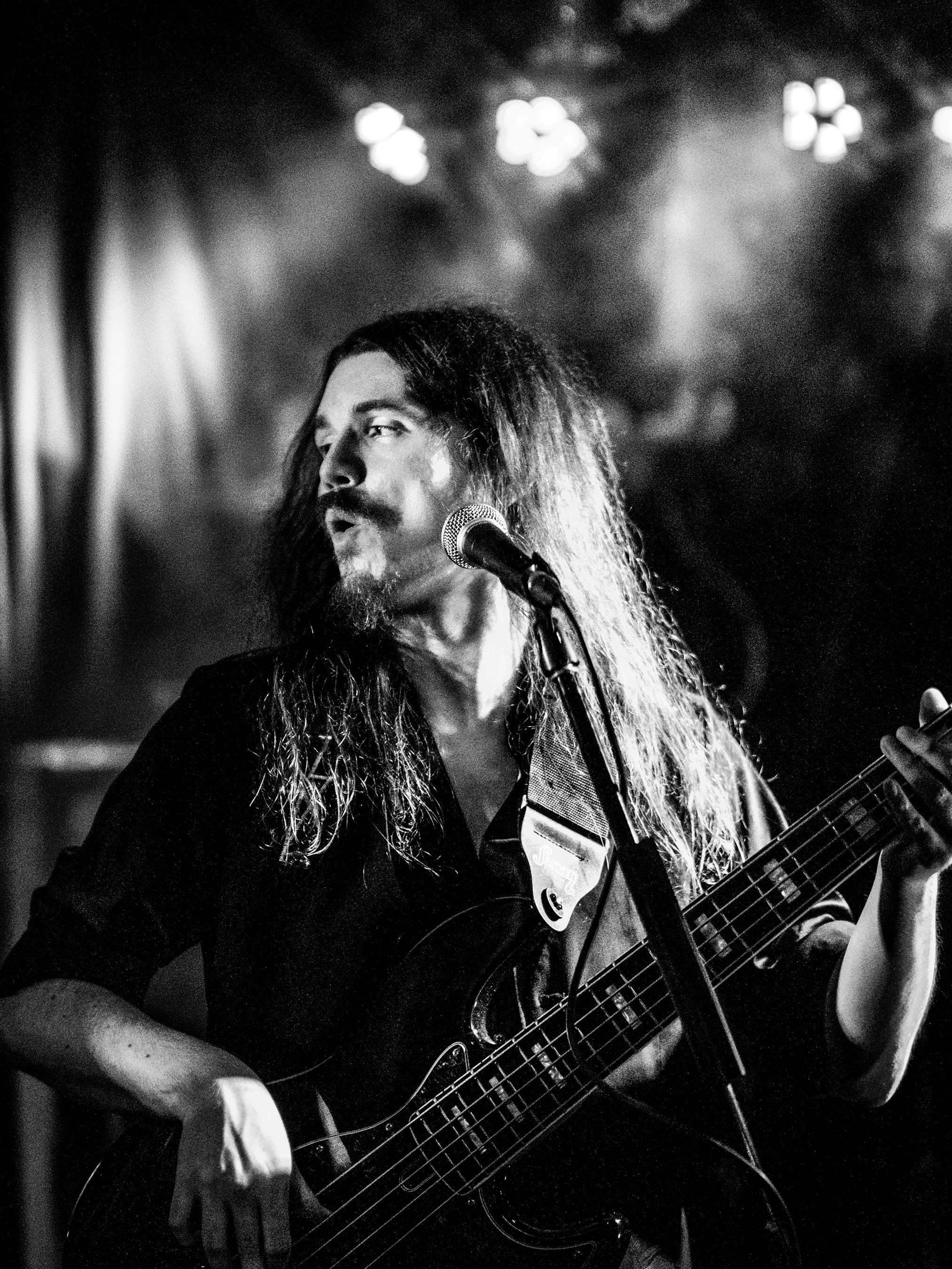 Black and white photo of a man with long hair and a beard playing an electric bass and singing into a microphone in a dimly lit setting.