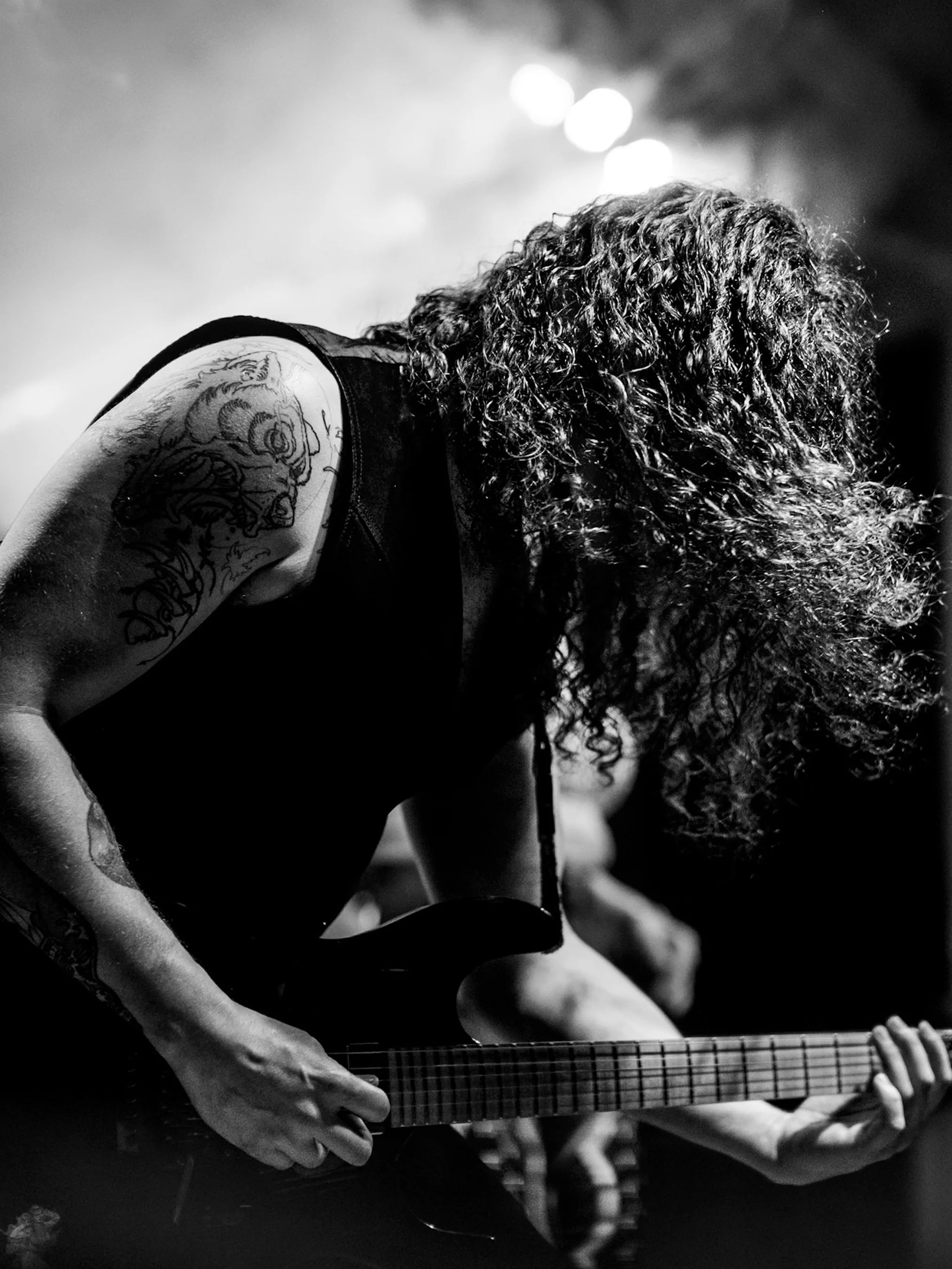 A person with long, curly hair playing an electric guitar, wearing a sleeveless shirt with a tattoo on the arm, in a dim setting with bright lights in the background.