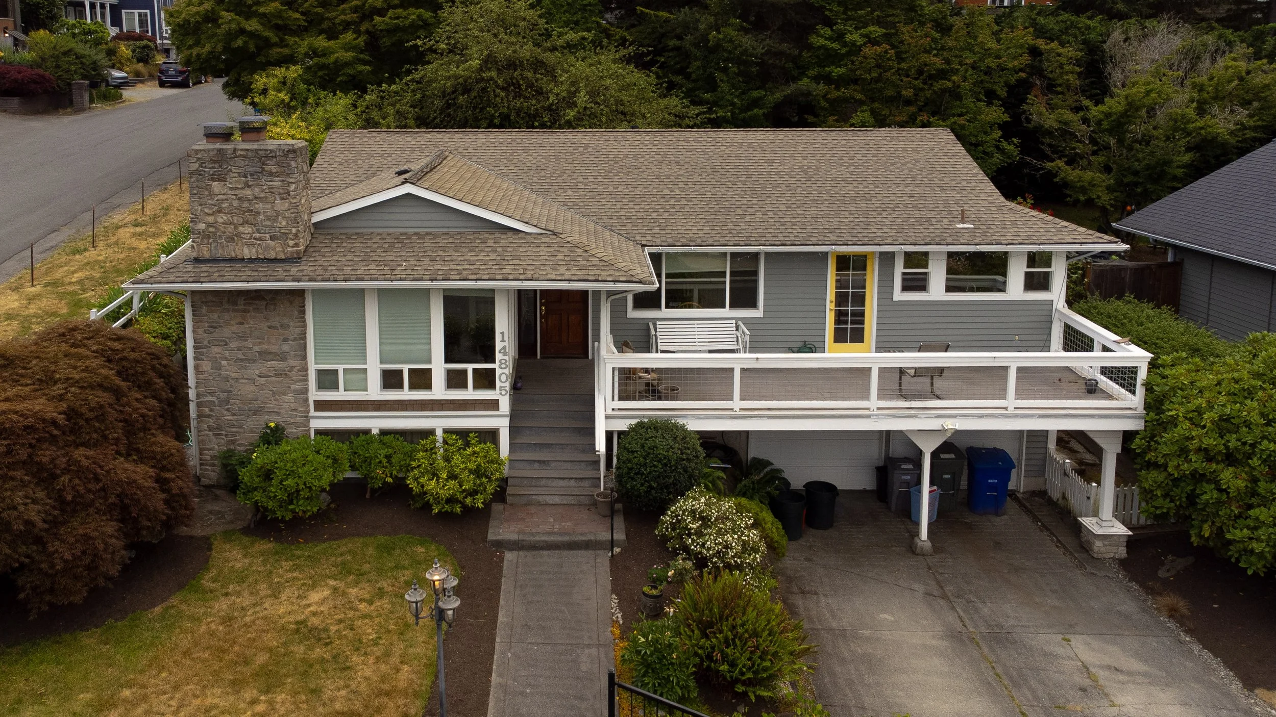 A two-story house with a front porch, gray siding, a chimney, and a balcony, surrounded by trees and landscaped yard.