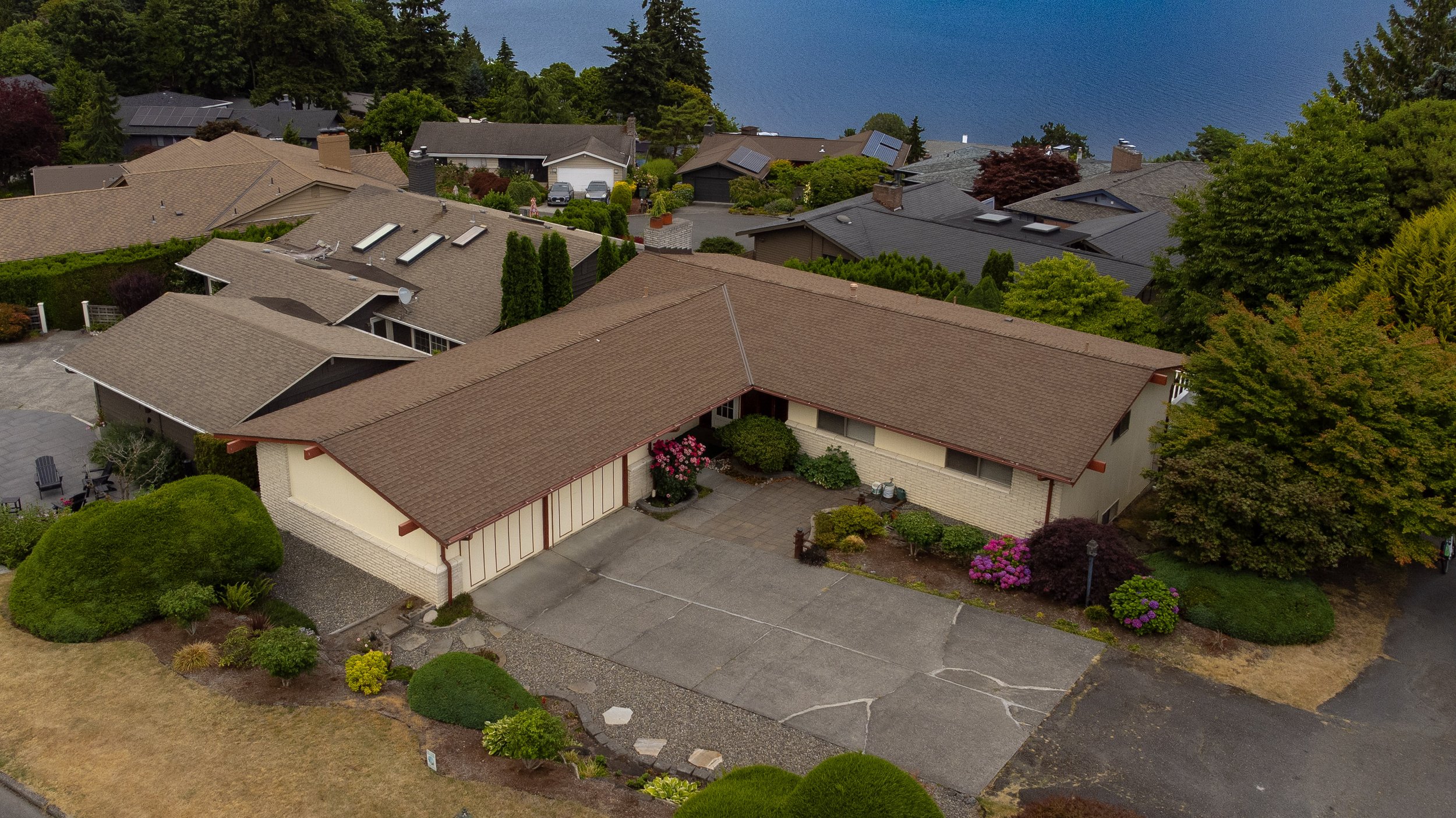 Aerial view of a suburban neighborhood with multiple houses featuring brown, gray, and dark roofs, surrounded by lush green trees, bushes, and landscaped gardens, near a body of water in the background.