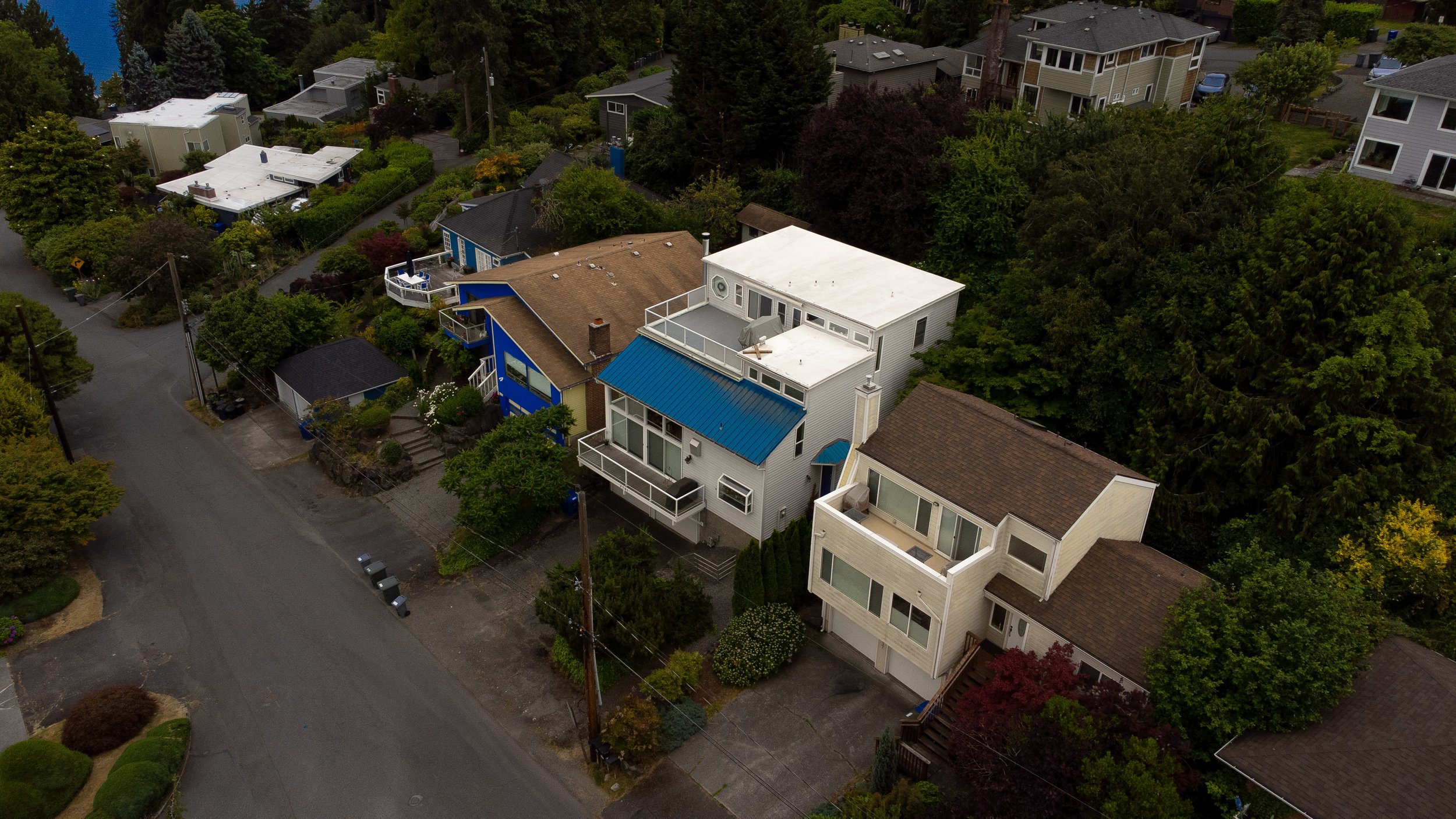 An aerial view of a residential neighborhood with multiple houses, surrounded by trees and greenery. The houses vary in size and color, with some featuring decks and flat roofs.
