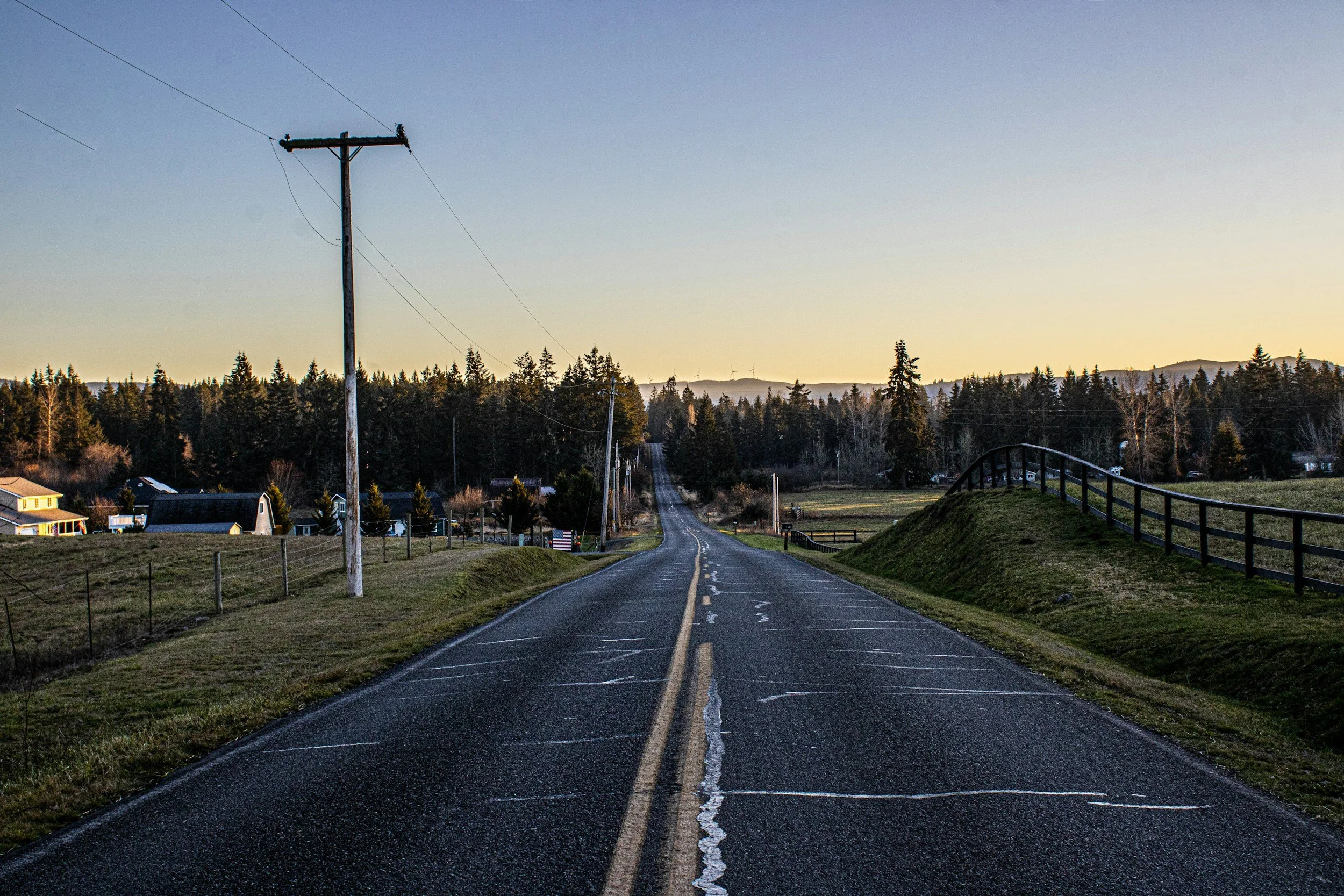 A rural road leading into a forested area with houses on the left and a wooden fence on the right, utility pole on the left, under a clear sky during sunset.