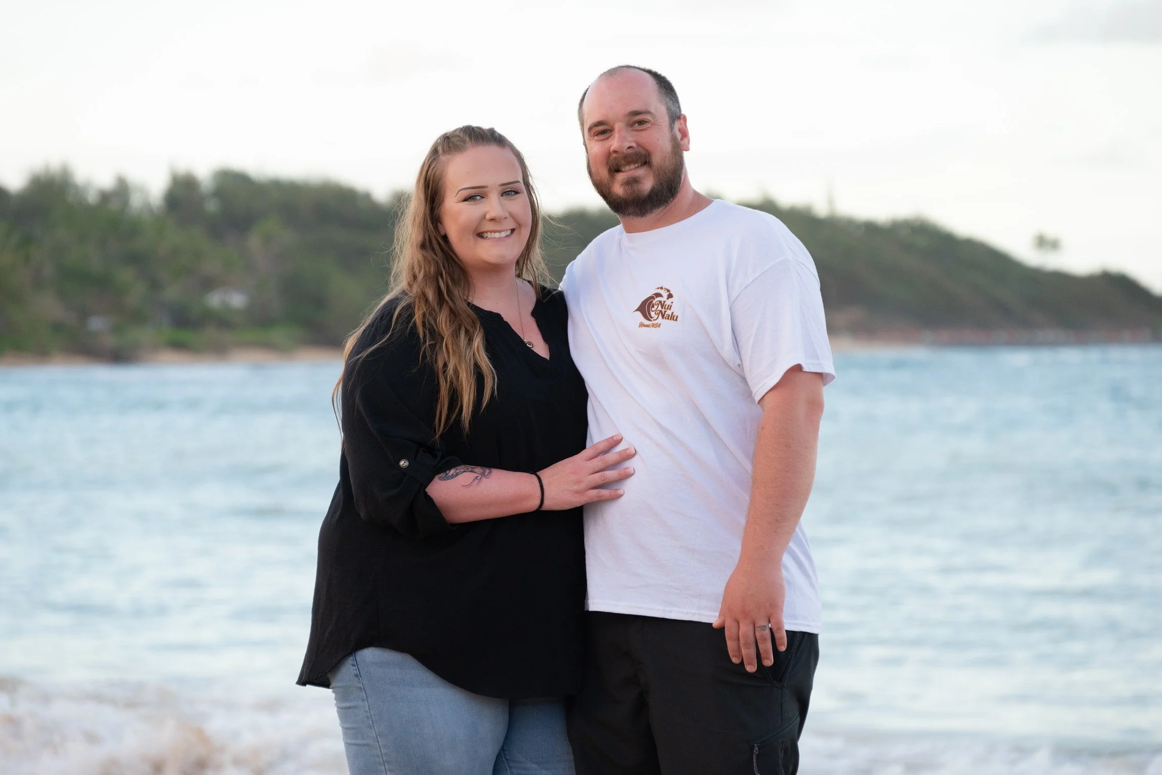 A smiling couple stands by the water on a beach, with the woman wearing a black top and jeans and the man wearing a white T-shirt and black pants.