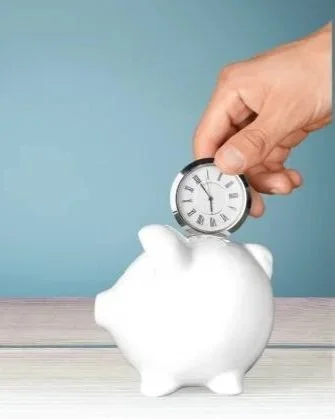 A hand placing a clock into a white piggy bank on a wooden surface with a blue background.