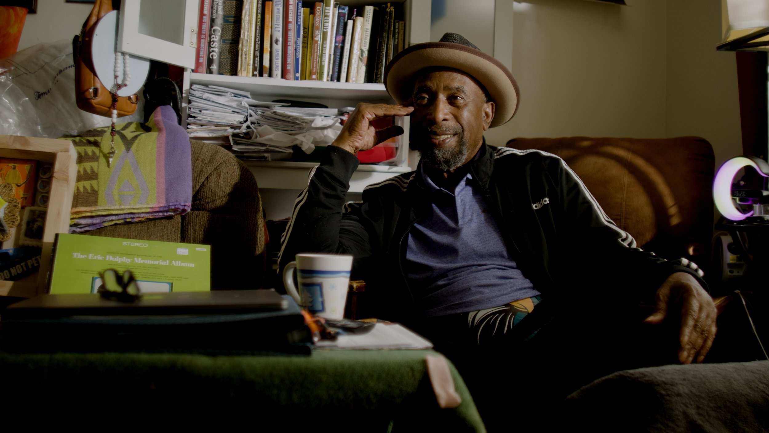 A man with a beard wearing a tan fedora hat and a black and white Adidas jacket is sitting on a couch, smiling, with his right hand raised to his temple, in a cluttered room with a bookshelf, papers, a colorful towel, and a lamp in the background.