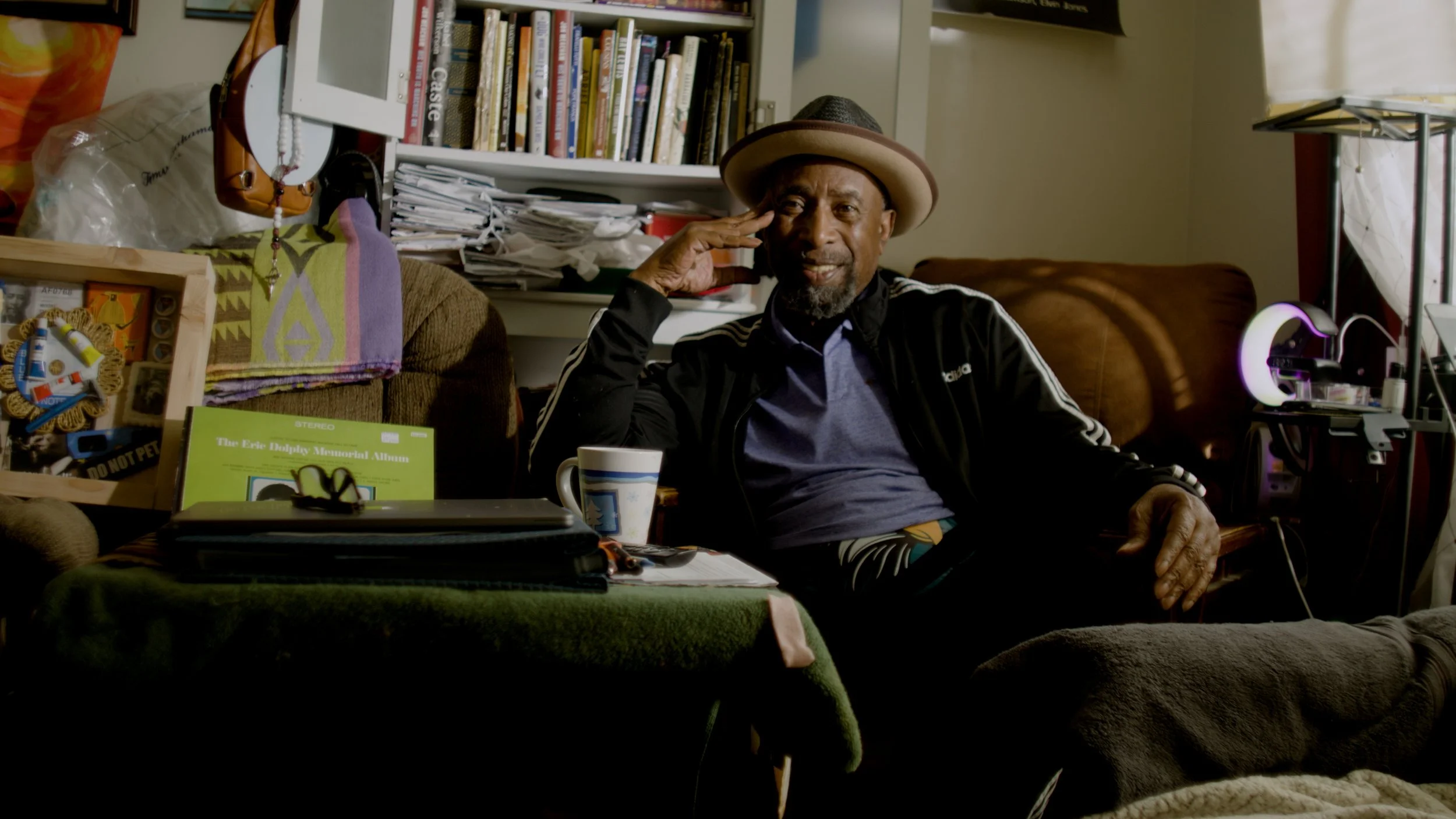 An older man with a beard and mustache wearing a brown hat, black jacket with white stripes, and a blue shirt, sits in a cluttered room with books, papers, and various objects. He is saluting with his right hand, smiling, and sitting on a brown couch