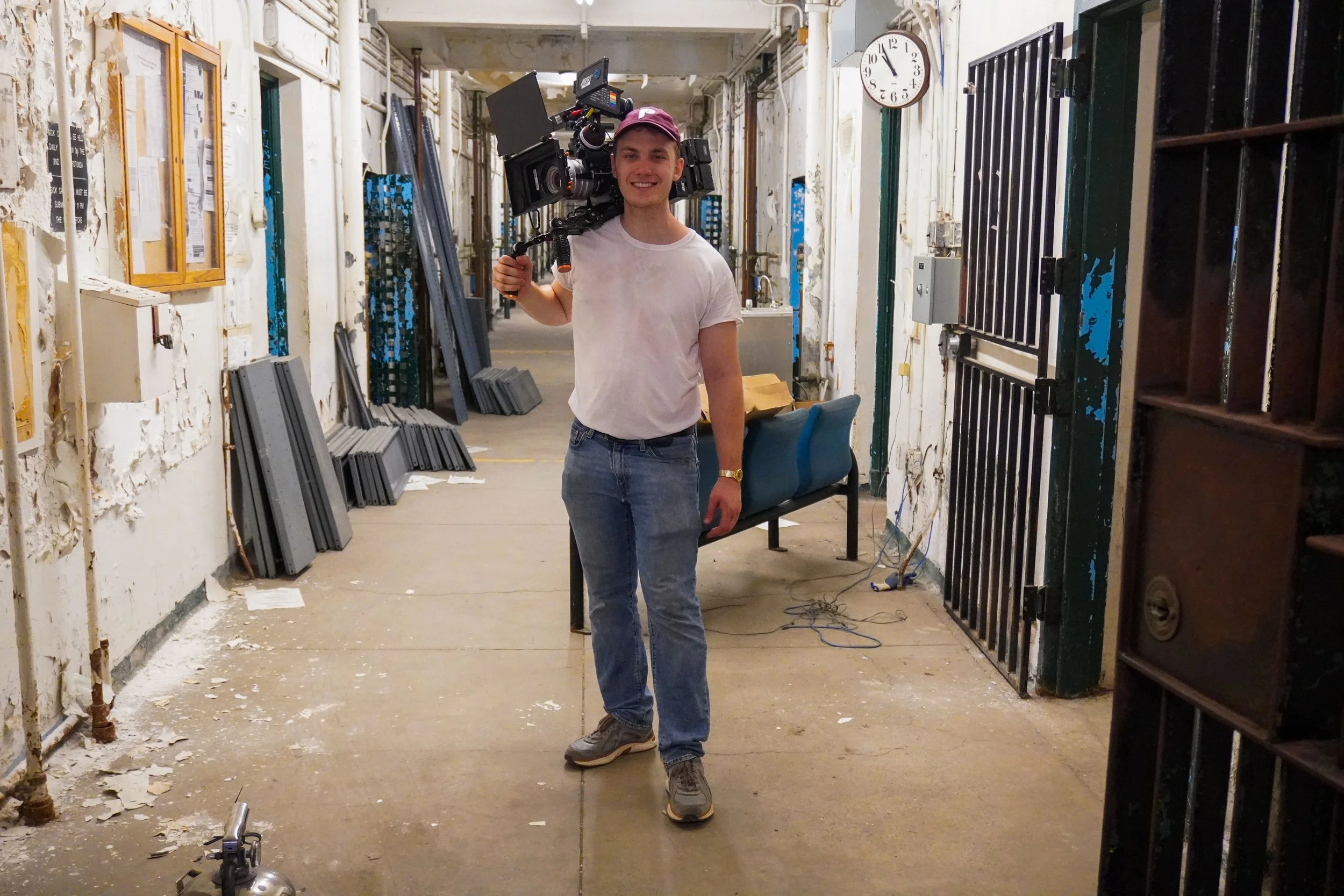 A young man in a pink cap and casual clothes smiling at the camera while carrying a professional video camera on his shoulder in a worn, industrial hallway with peeling paint and equipment.
