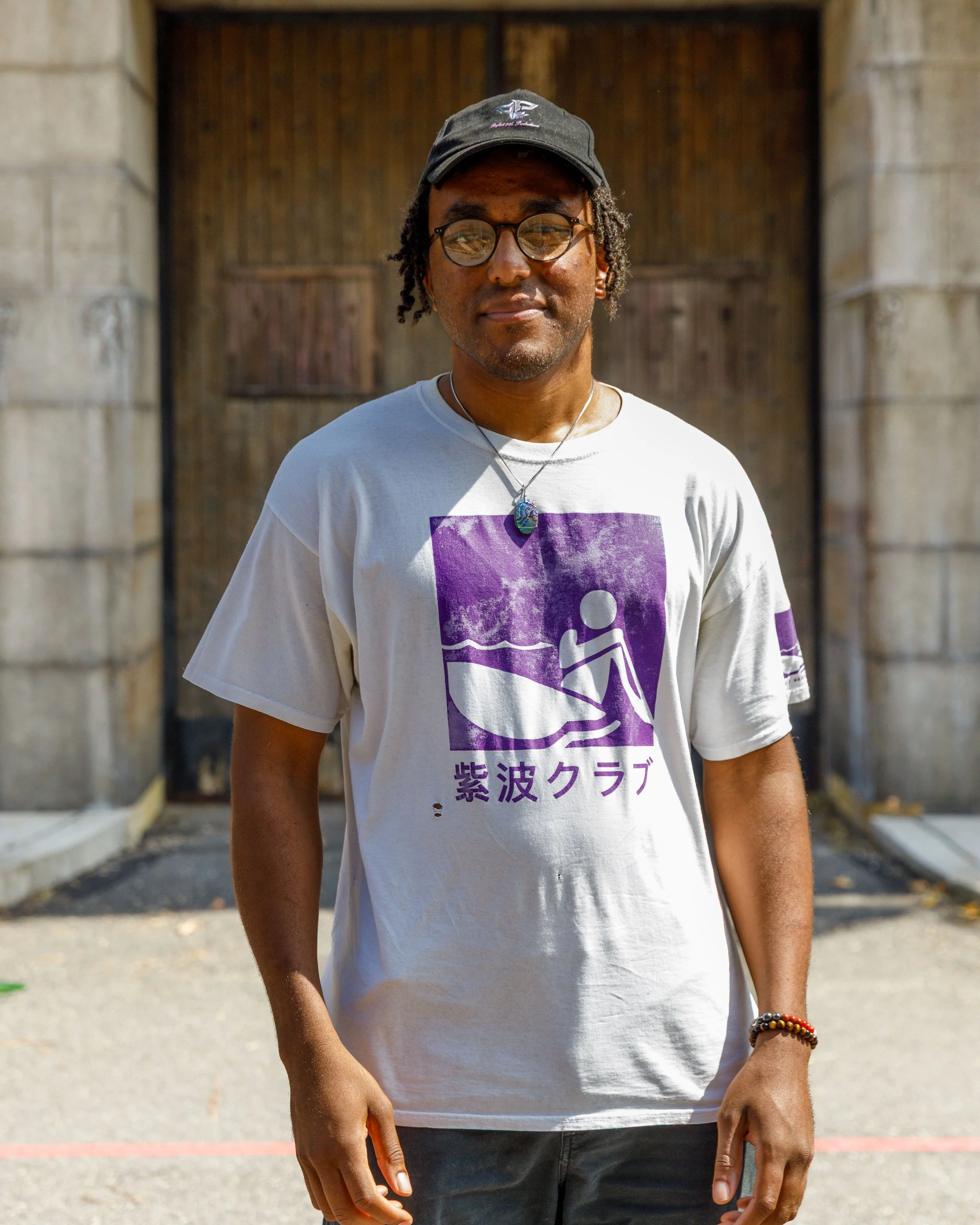 A man with dark curly hair and glasses, wearing a black cap, white graphic t-shirt with purple and black design and Japanese characters, stands outdoors in front of a wooden door framed by stone columns, with his right arm bent and hand relaxed at his side.