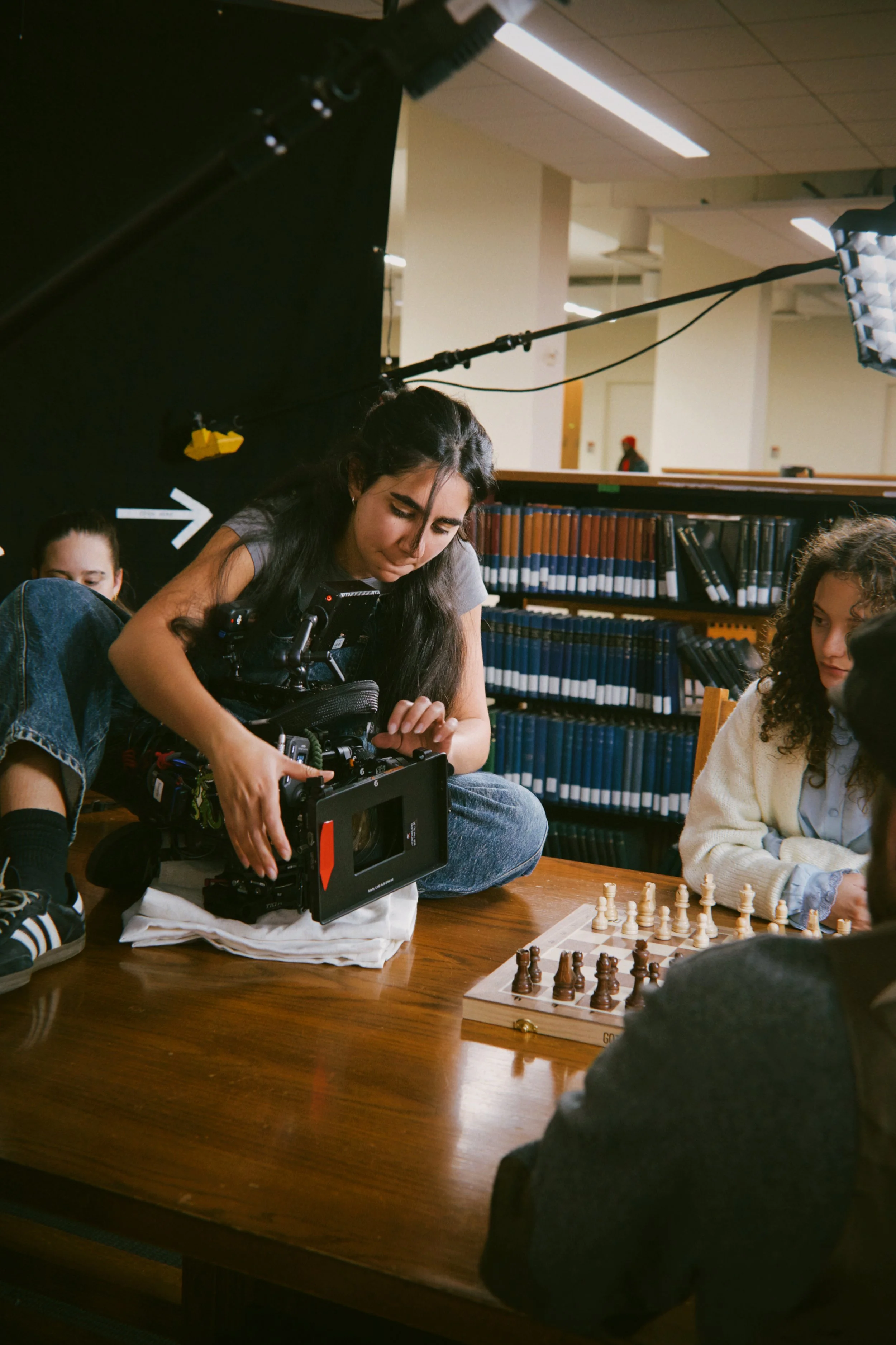 A woman operating a professional camera while sitting on the floor at a table during a chess game, with children and bookshelves in the background.