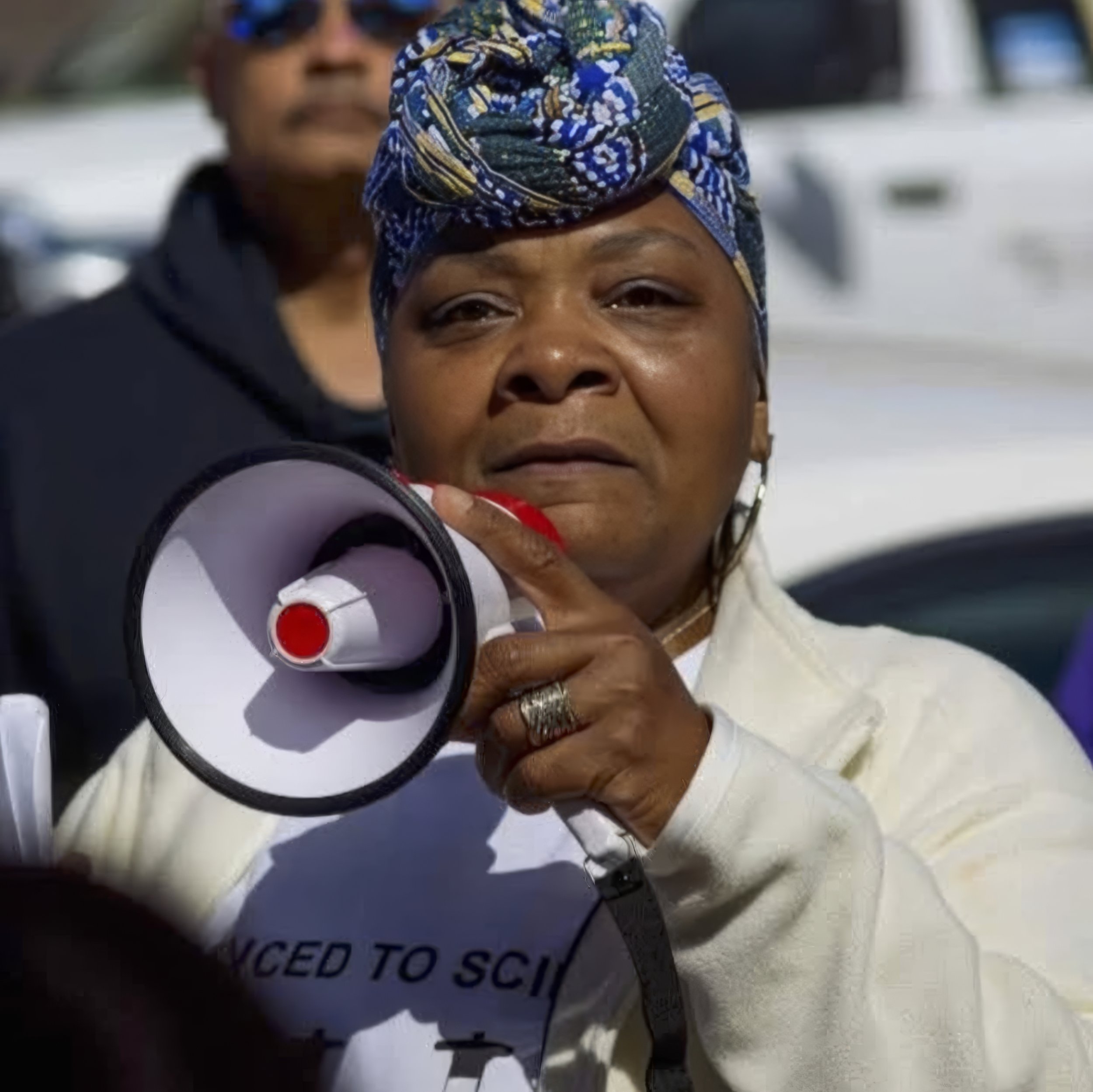 Close-up of a woman with dark skin wearing a blue patterned headwrap and a white jacket, holding a megaphone. Behind her, a person wearing sunglasses and a dark jacket is visible, along with vehicles in the background.