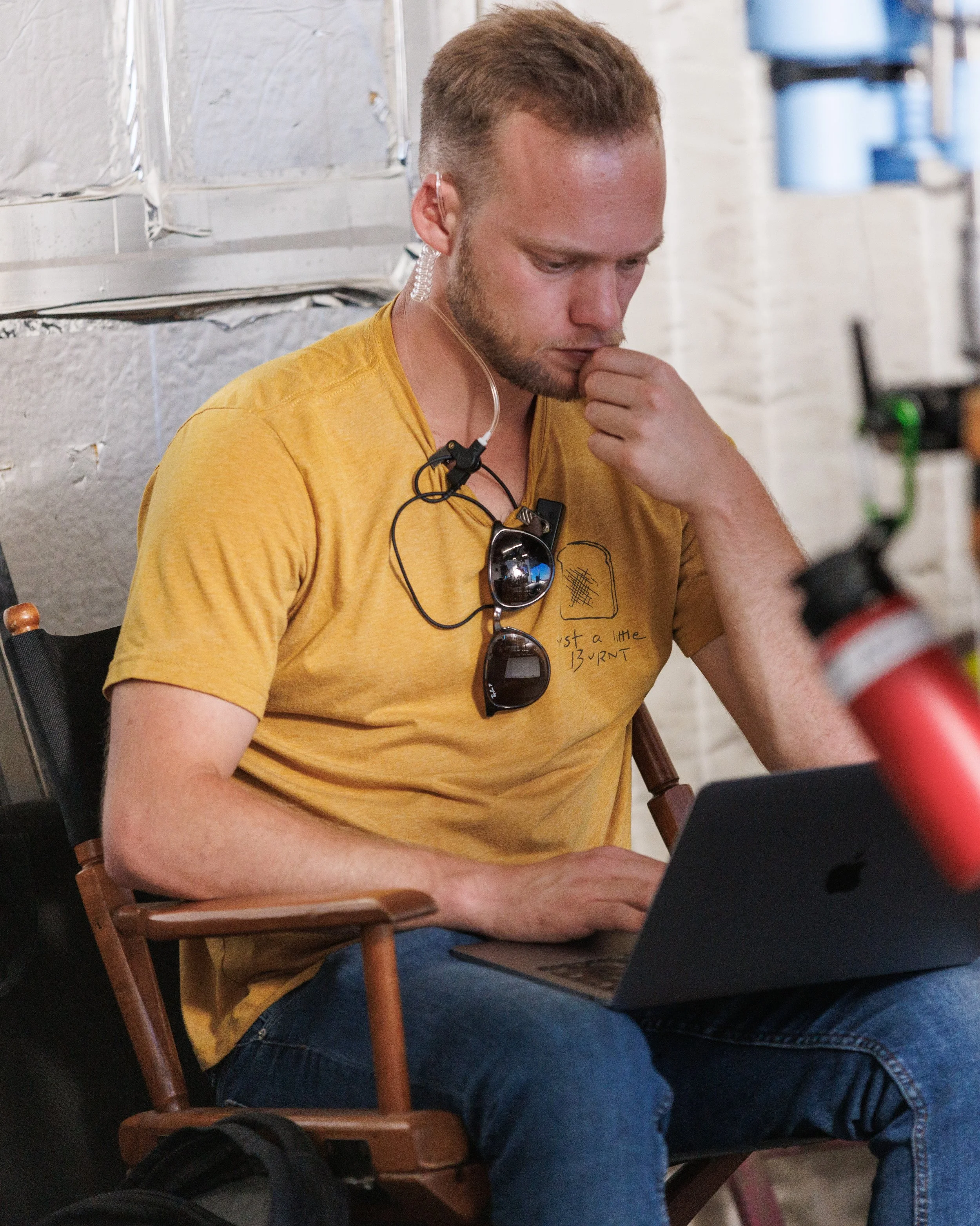 A man with short brown hair, a beard, and a yellow t-shirt sitting on a wooden chair, looking at a laptop on his lap, with glasses hanging from his shirt's neckline, and a person in the foreground with a red wristband.
