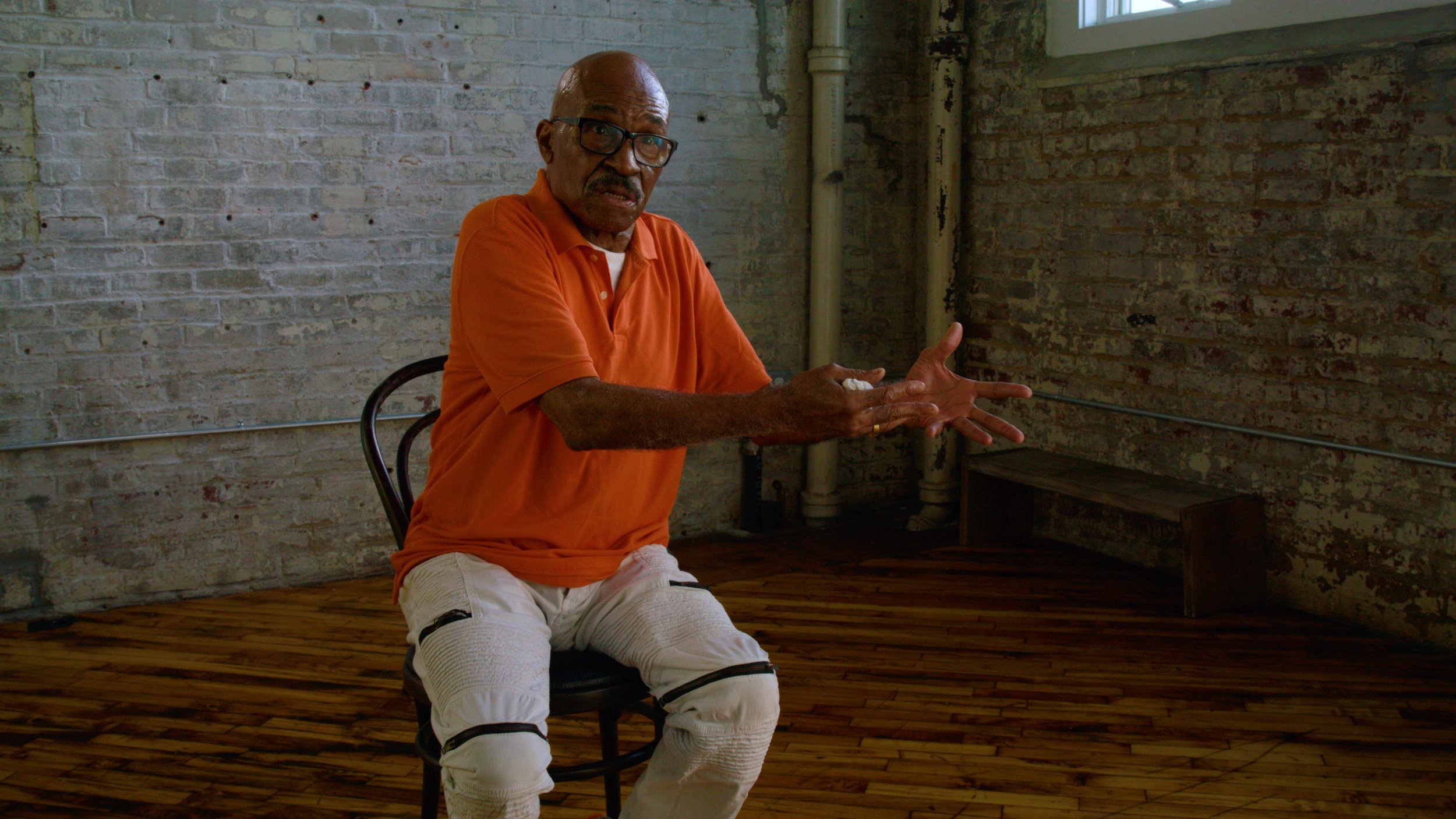 An older man with glasses, wearing an orange polo shirt and white knickers, sitting on a chair in a room with brick walls and wooden floors, gesturing with his hands.