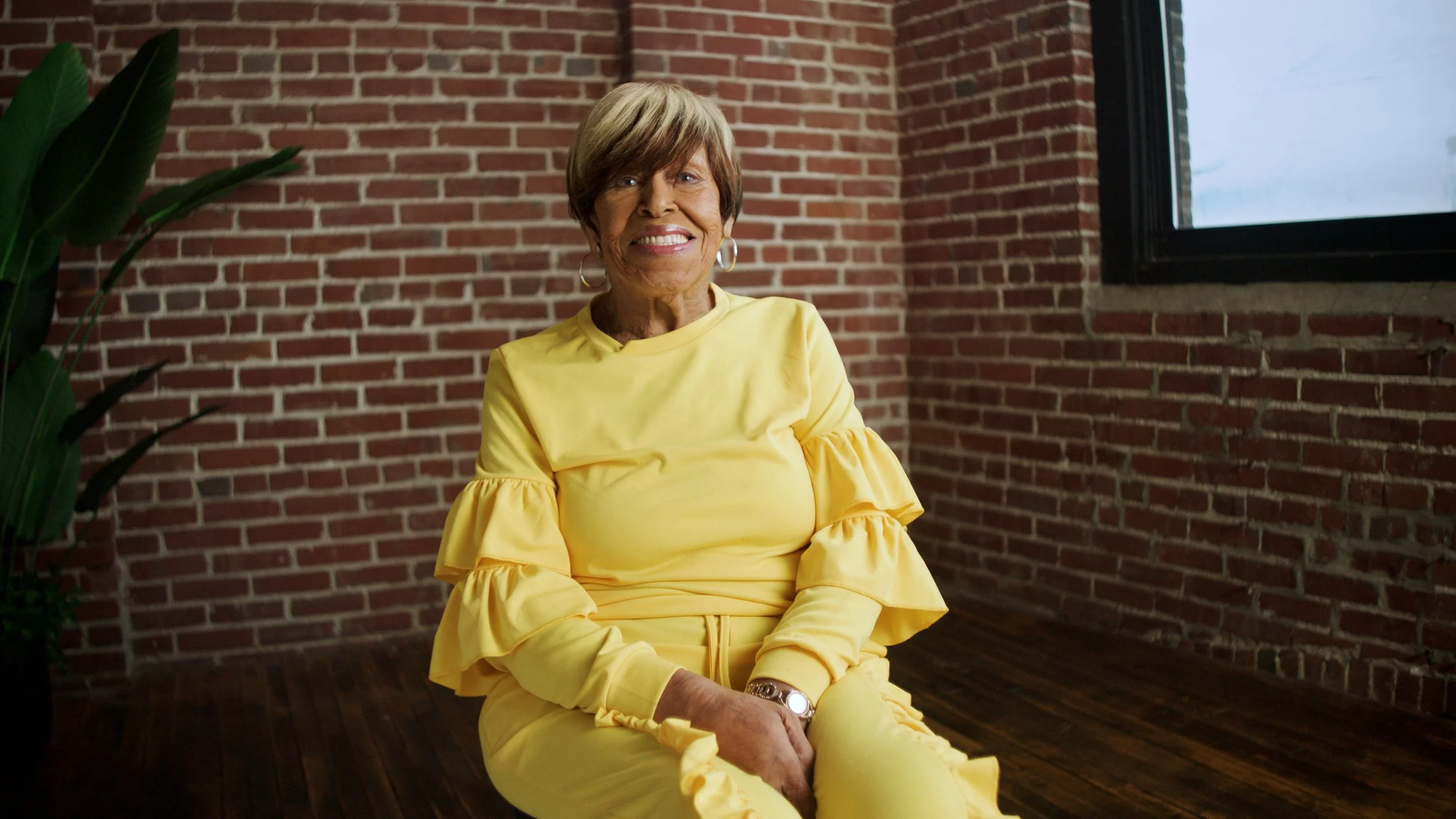 An elderly woman with short hair smiling, seated in a room with brick walls and a large window, wearing a yellow dress with ruffled sleeves.