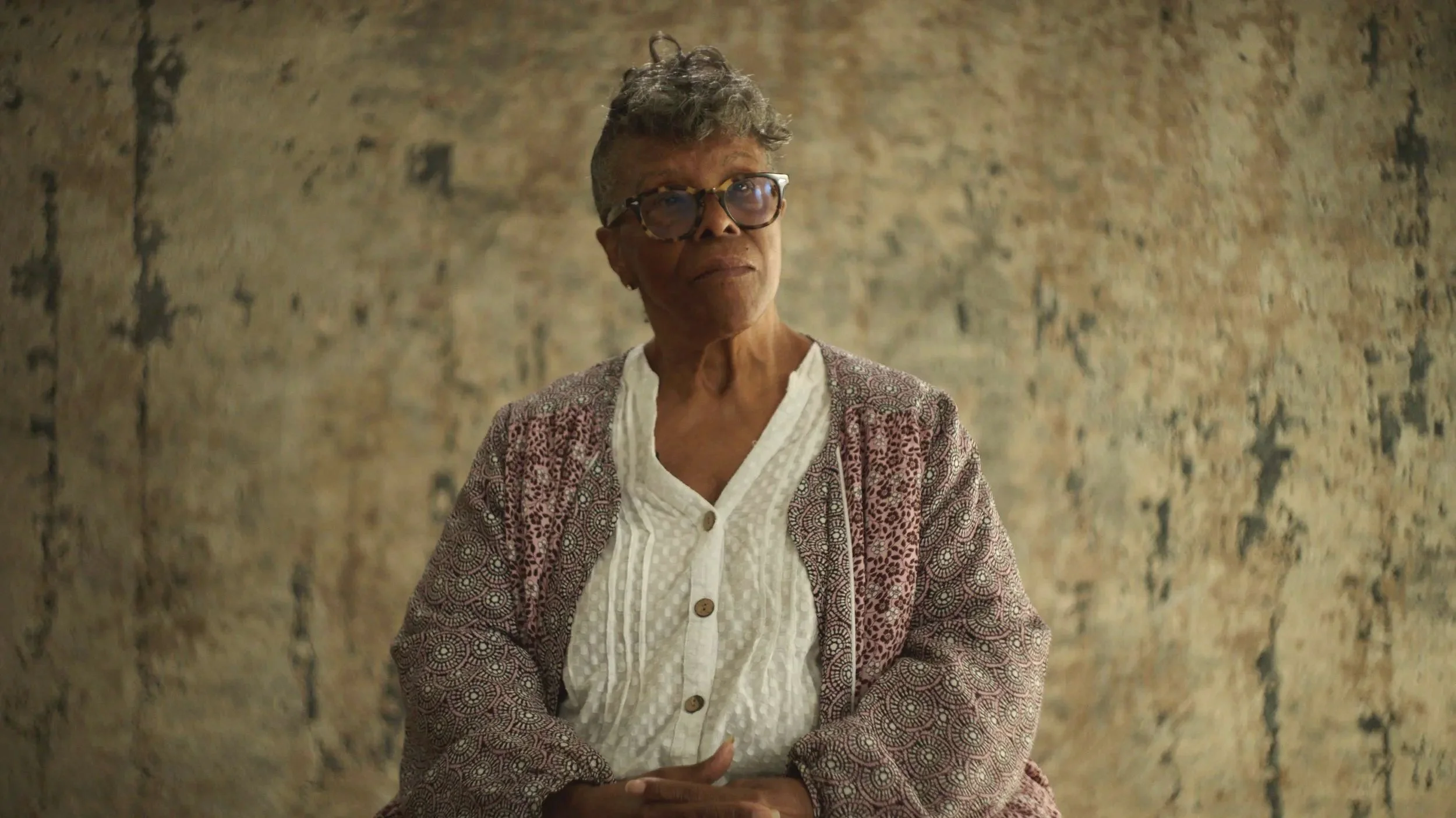 An elderly woman with short, curly gray hair and glasses looks slightly upward with a thoughtful expression. She is wearing a white blouse and a patterned cardigan, standing in front of a textured, beige wall.