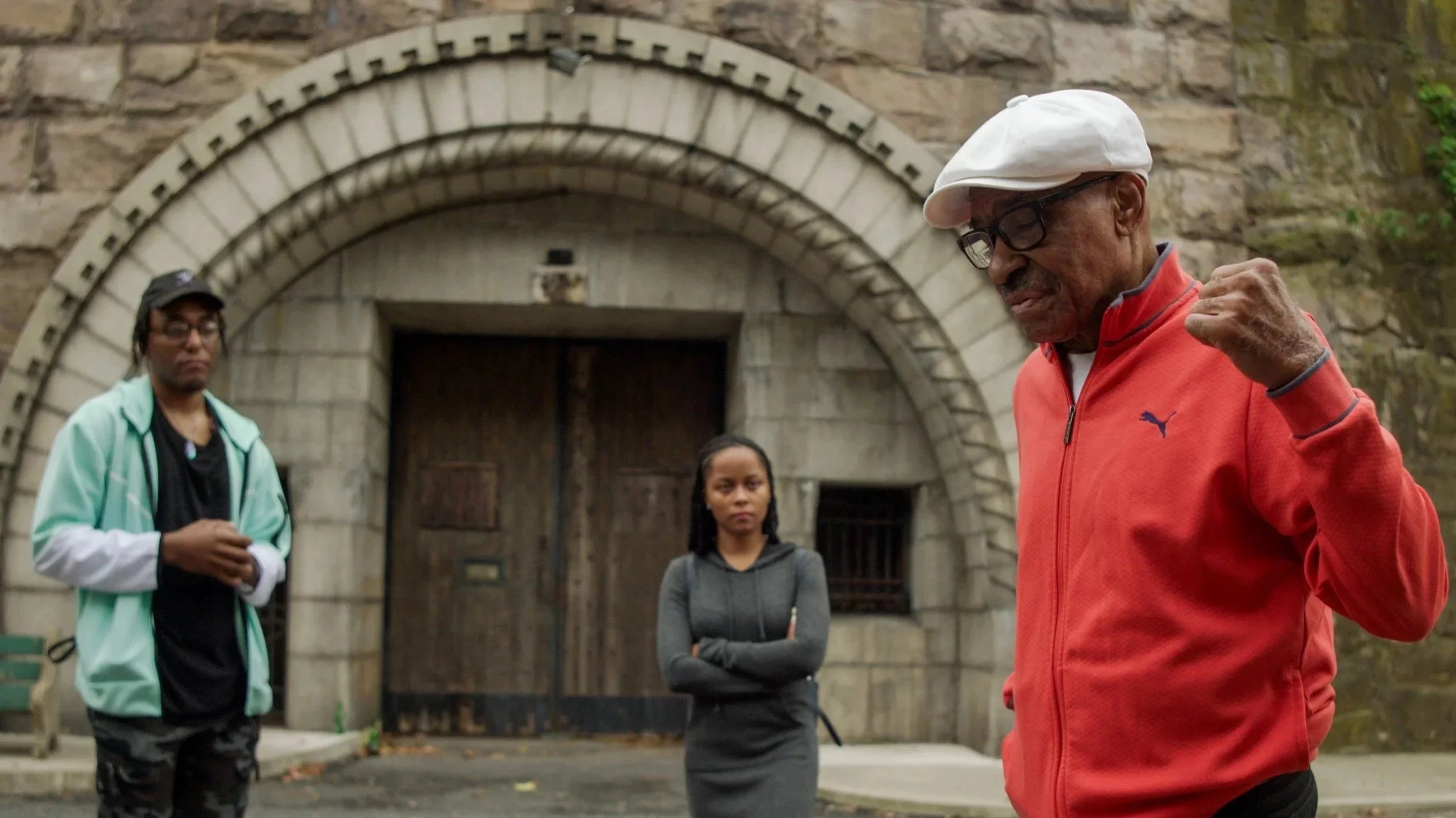 Four people standing outdoors in front of a stone archway. An elderly man wearing glasses, a white cap, and a red jacket is speaking with a fist raised. Two younger women and one younger man listen, with the women appearing attentive and the man look