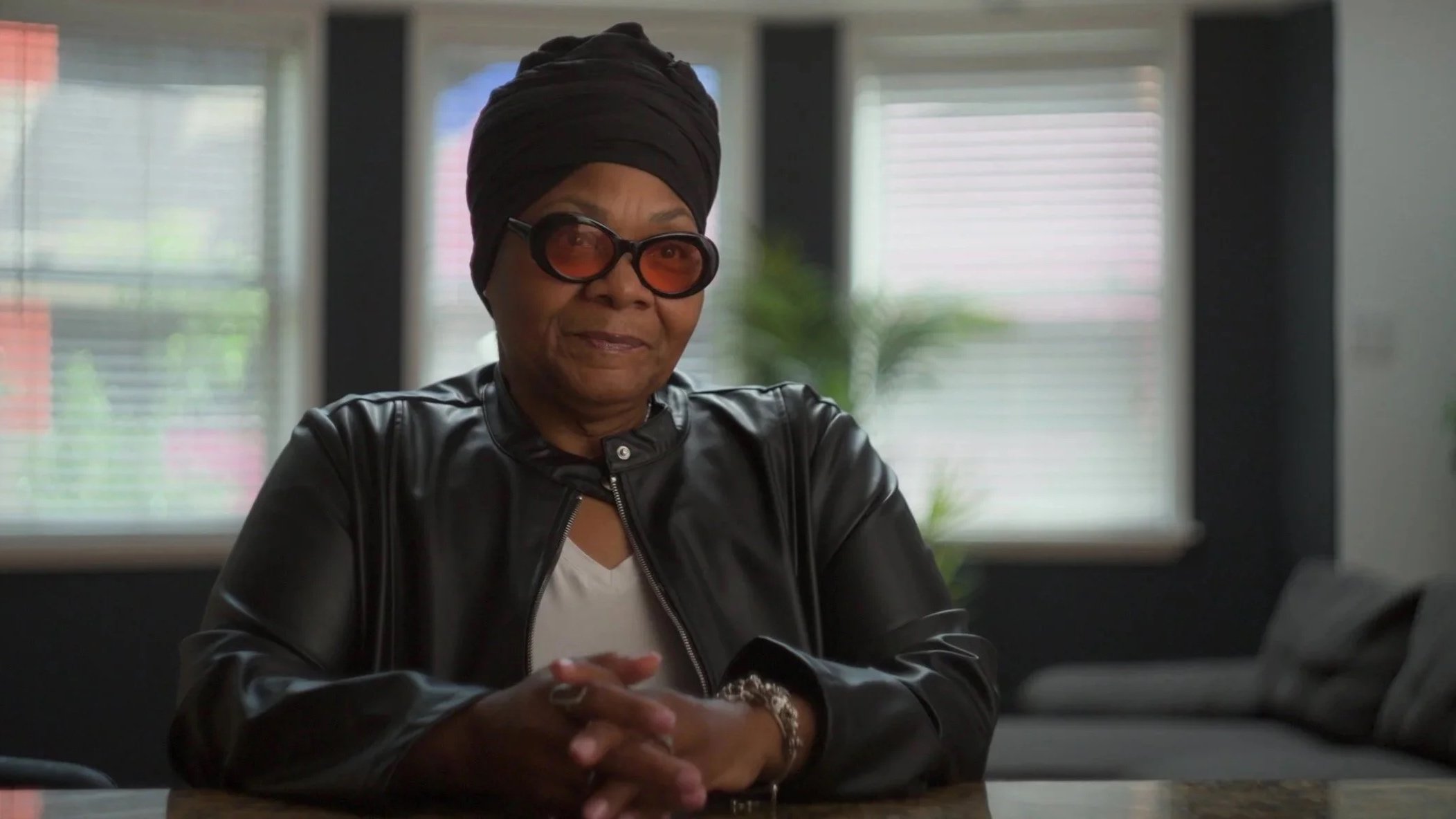 A woman wearing a black headwrap, oversized sunglasses, a black leather jacket, and a white shirt, sitting at a table in a room with large windows and blinds.