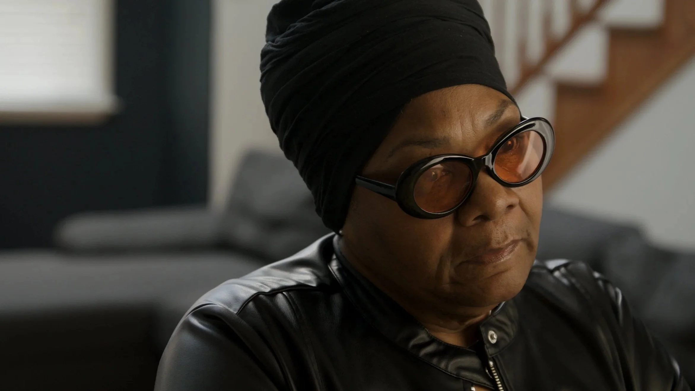 Close-up of an African American woman wearing large round glasses, black head wrap, and leather jacket, looking focused.