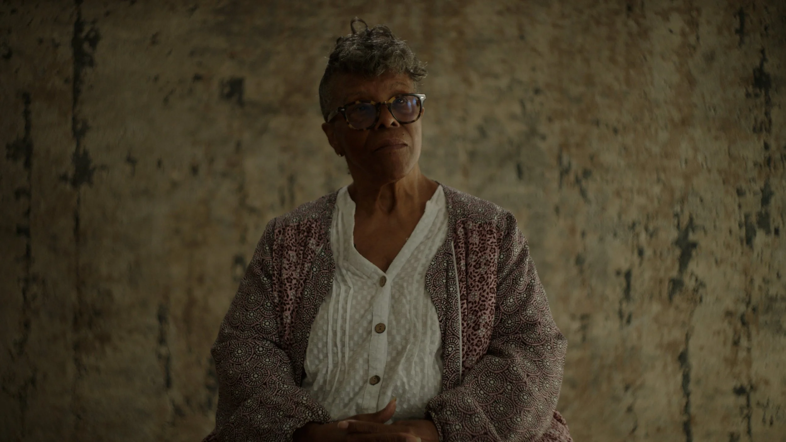 An elderly woman wearing glasses, a white blouse, and a patterned cardigan, standing against a textured wall.