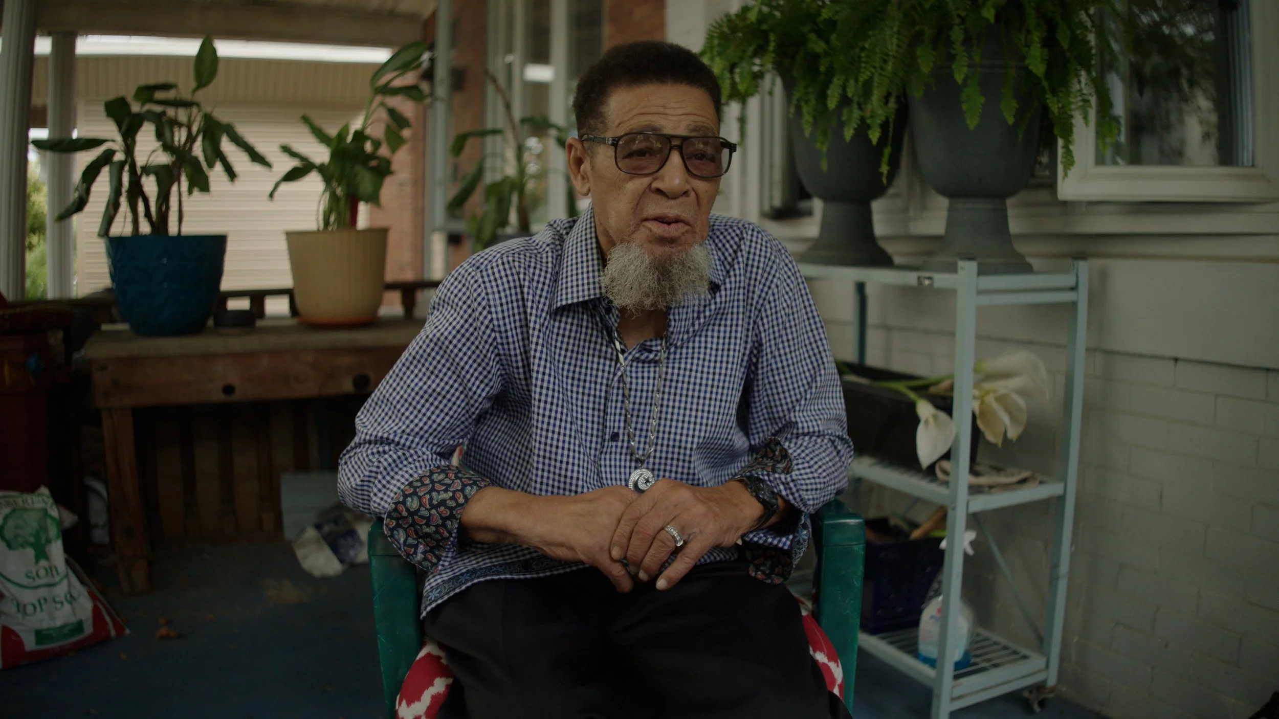 An older man with glasses and a beard sitting in a porch area surrounded by potted plants.