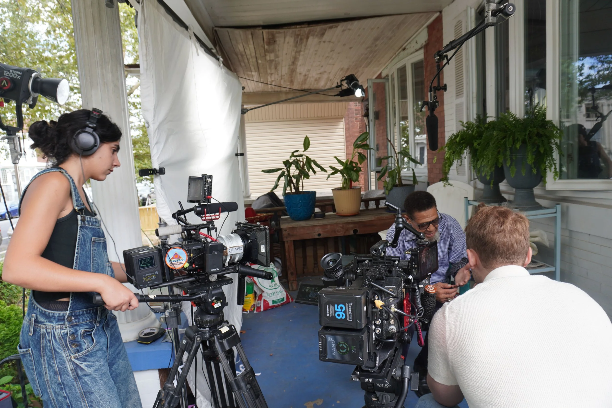 People working on a film set, with cameras and sound equipment outdoors on a porch, with plants and trees in the background.