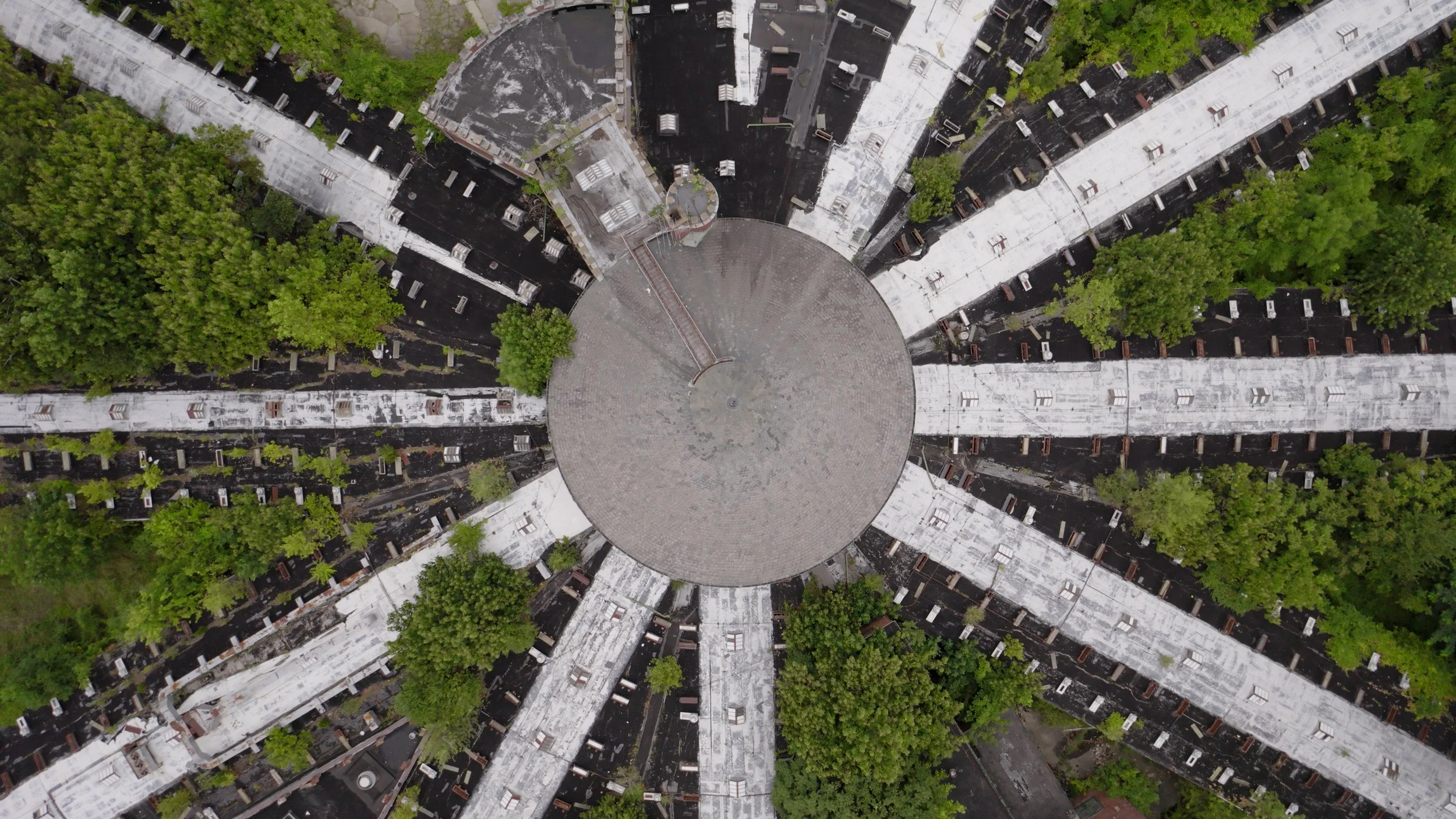Aerial view of a circular plaza with radiating walkways, surrounded by lush green trees and black rooftops.