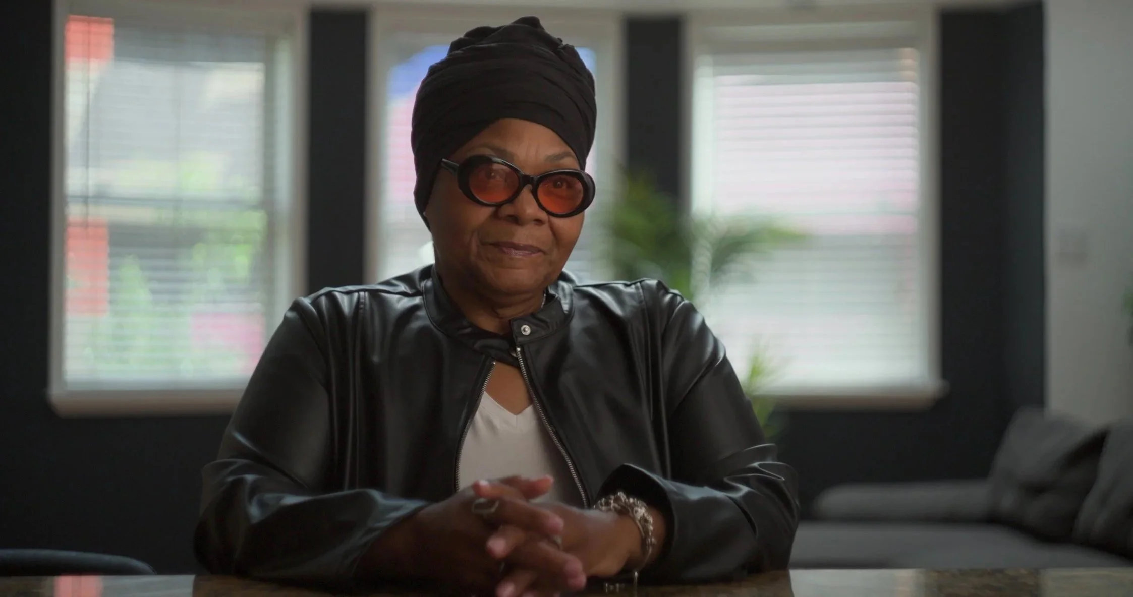 A woman wearing a black headwrap, sunglasses, and a black leather jacket sits at a table in a room with windows behind her.